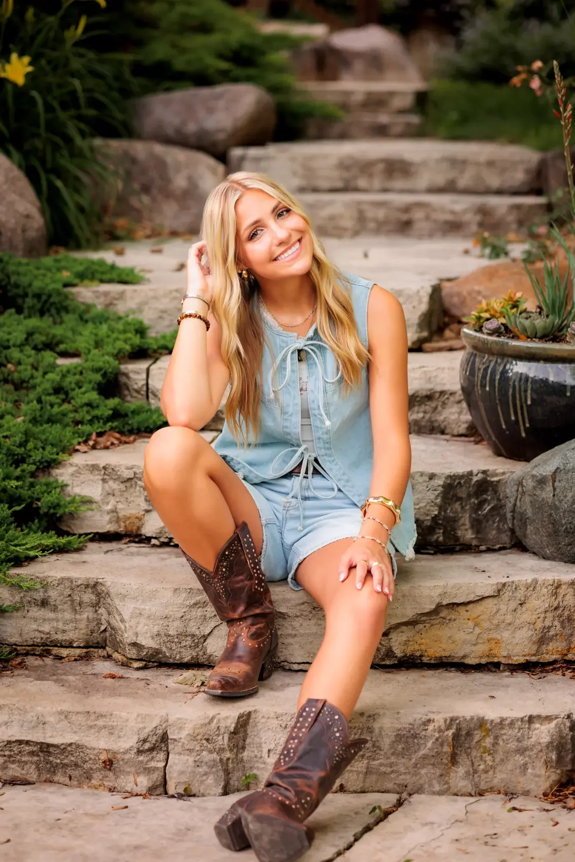 High school senior girl with long blonde hair, wearing a light blue sleeveless top, denim shorts, and brown cowboy boots, sitting on stone steps outdoors surrounded by green plants and rocks, smiling at the camera.