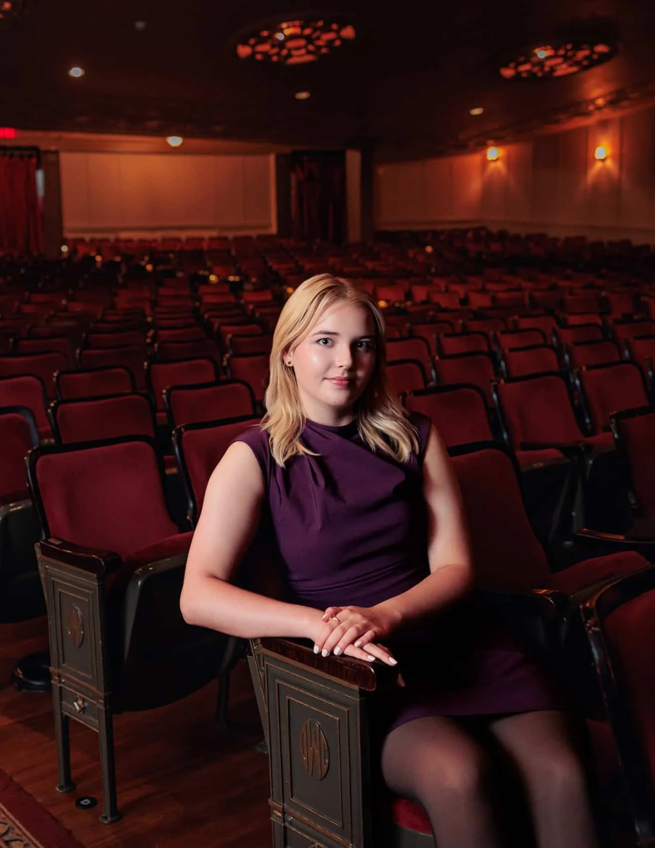 Senior girl with blonde hair sitting alone in an empty theater auditorium with red seats at Hoyt Sherman Place in Des Moines, wearing a dark purple dress.