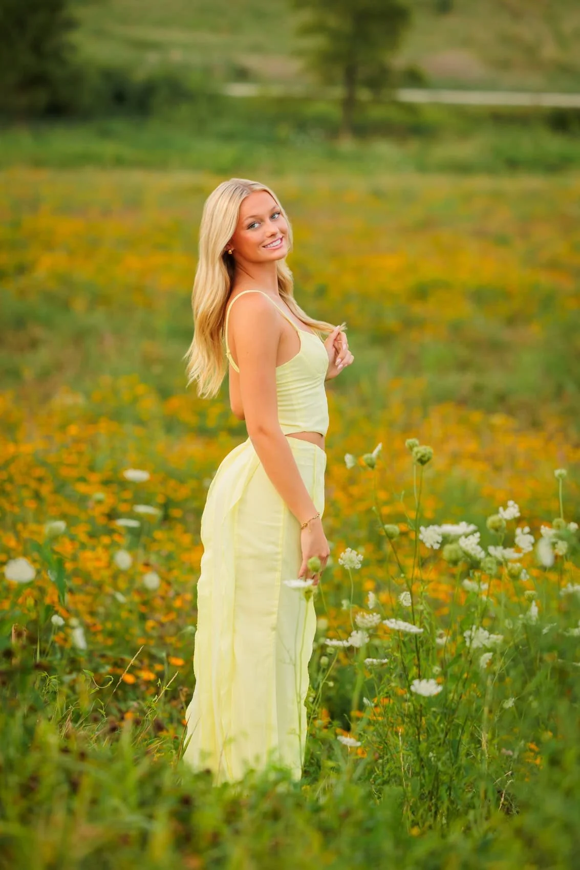 A young woman with long blonde hair in a yellow crop top and matching long skirt standing in a field of wildflowers, smiling at the camera.