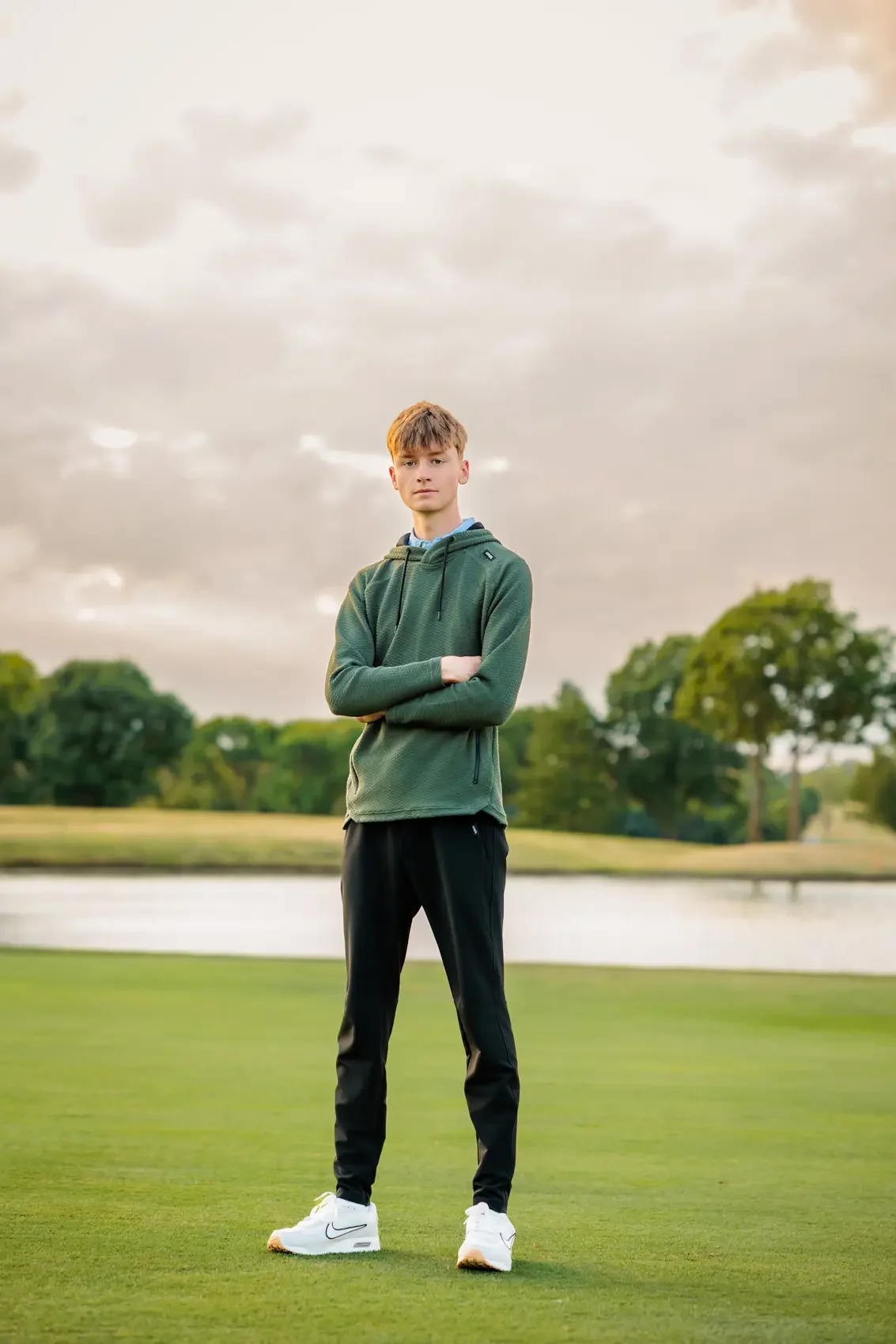 Senior boy standing on a grassy area near a body of water on a golf course in Des Moines with trees in the background. He is wearing a green hoodie, black pants, and white sneakers, with his arms crossed.