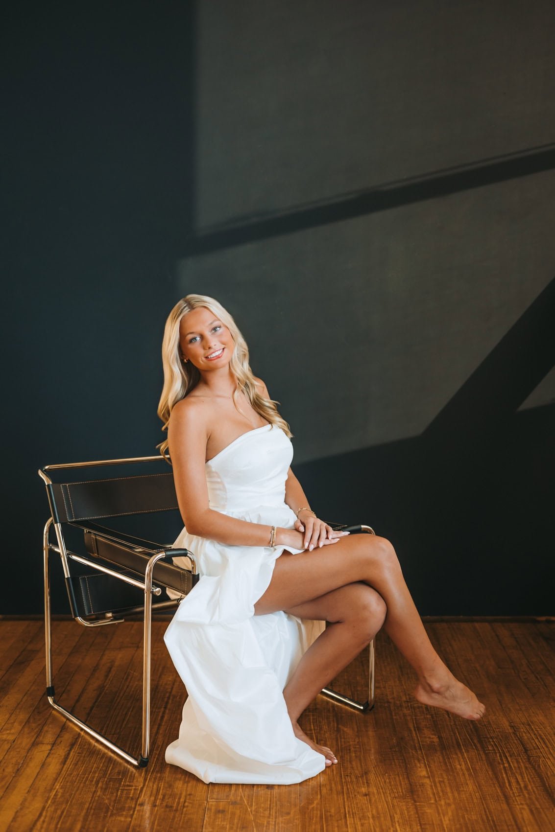 A young woman with long blonde hair sitting on a modern black and chrome chair on a wooden floor against a dark wall with geometric shadow patterns. She is wearing a white strapless dress and smiling.
