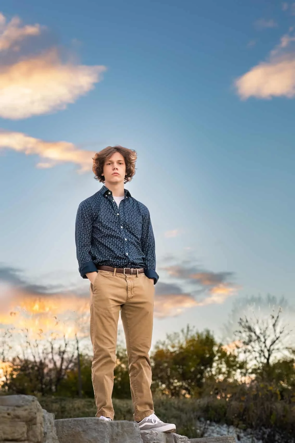  High school senior boy with curly hair stands on a rock outdoors during sunset, wearing a patterned blue shirt and beige pants, with trees and a colorful sky in the background.