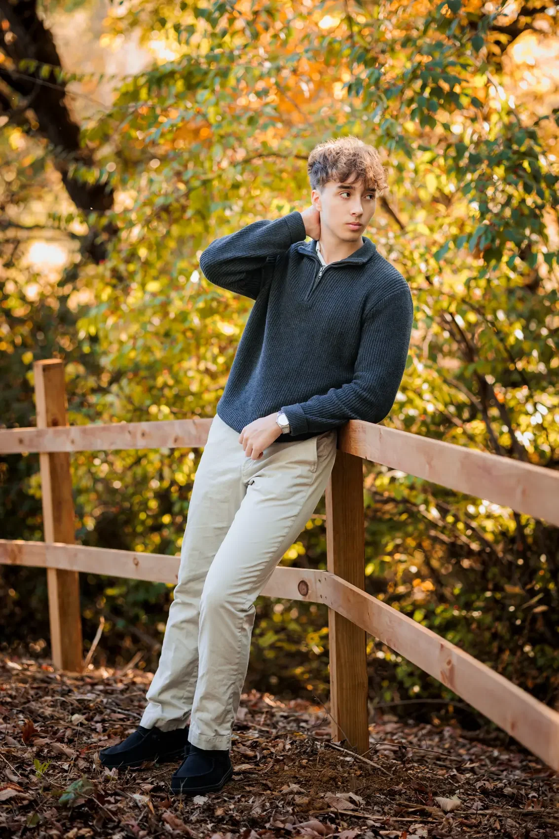 Senior boy standing outdoors in Des Moines, leaning against a wooden fence, with autumn leaves in the background. He is wearing a dark sweater and light-colored pants.