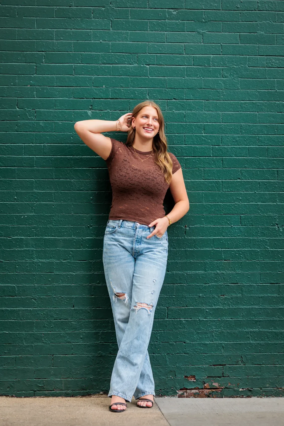 Senior girl with light brown hair wearing a brown lace top and ripped jeans, standing against a dark green brick wall. She is smiling and has her right hand behind her head.