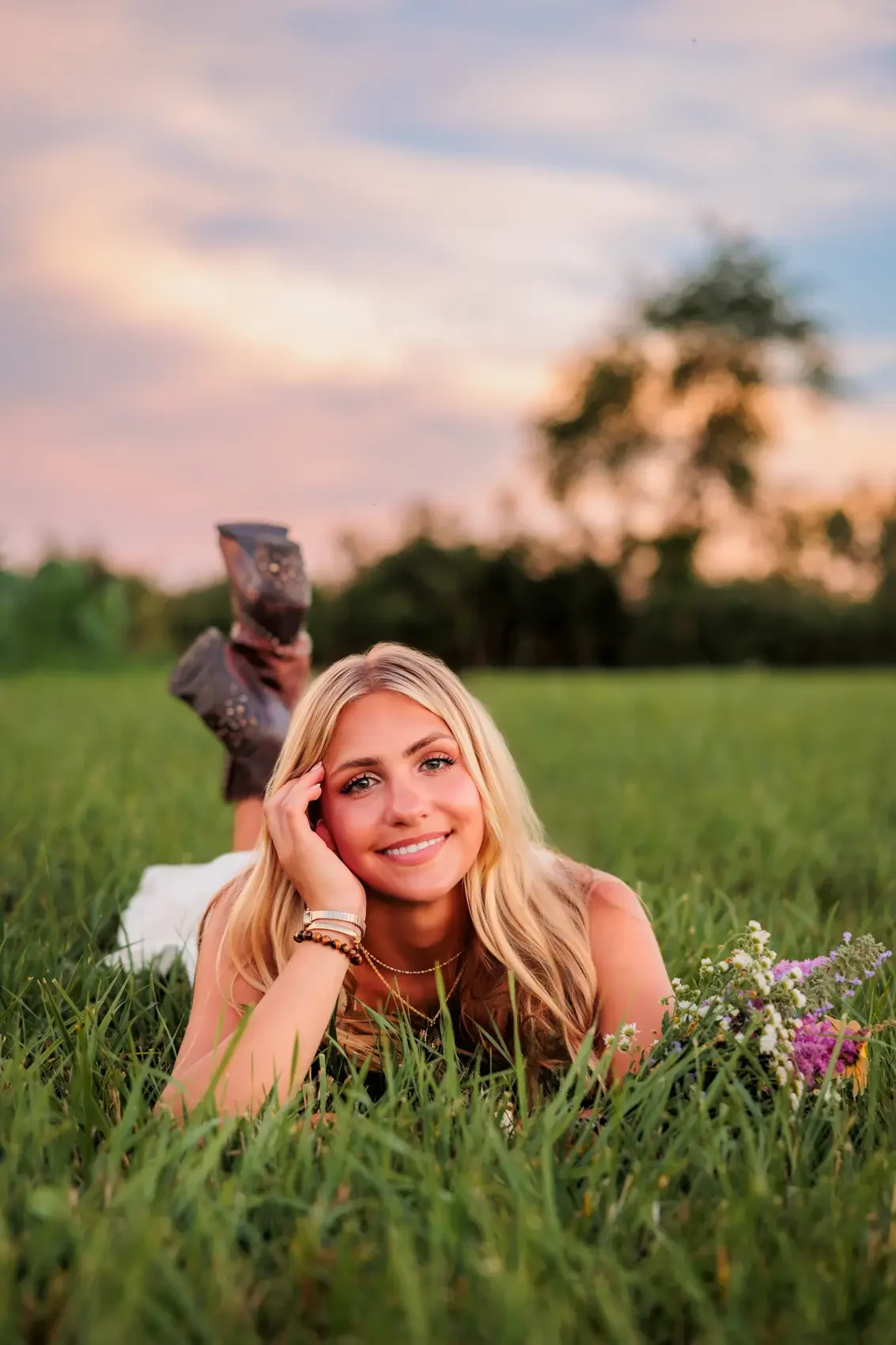Senior girl with blonde hair lying on her stomach on green grass, smiling at the camera during sunset, with trees and a colorful sky in the background.