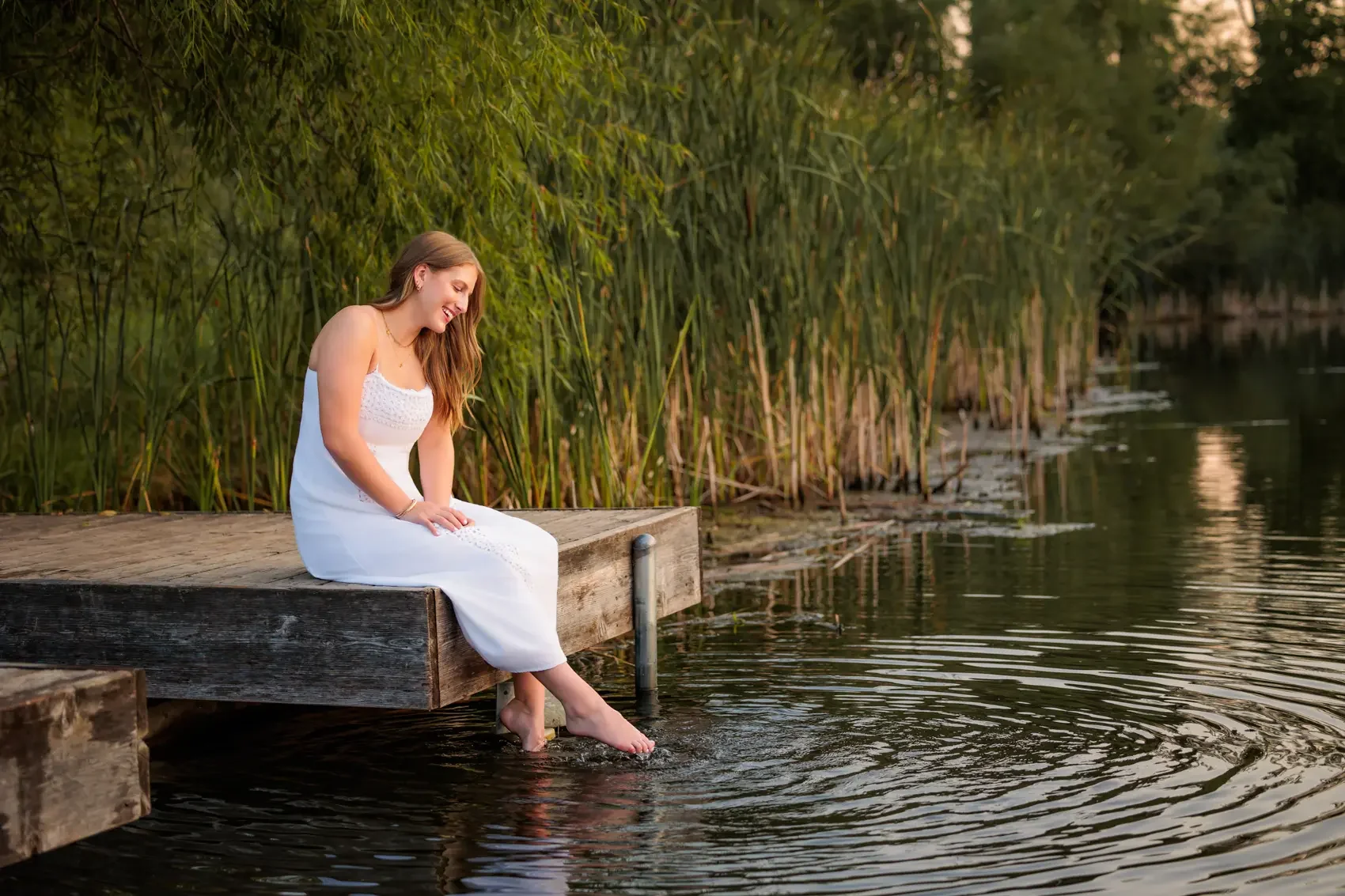 Senior girl  in a white dress sitting on a wooden dock with her feet in the water, smiling and looking down at the water near a lakeshore with tall green reeds in Des Moines.