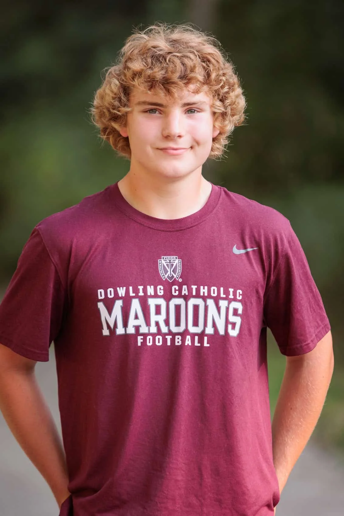  High school senior boy with curly blonde hair in a maroon 'Dowling Catholic Maroons Football' T-shirt standing outdoors with hands on hips, blurred background.