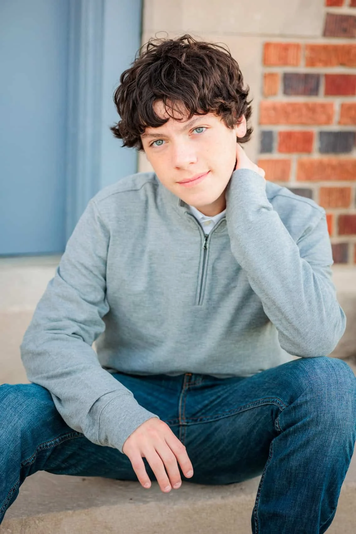 Senior boy with curly brown hair and blue eyes, wearing a light gray zip-up hoodie and blue jeans, sitting on the ground with legs spread and one hand resting on his neck, in front of a brick wall and blue door.