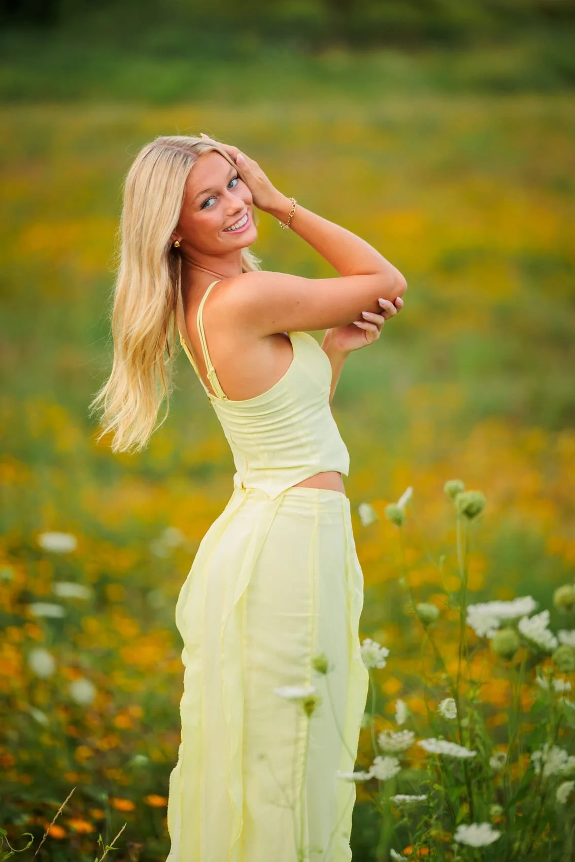 A smiling senior girl with long blonde hair, wearing a light yellow dress, standing in a field of flowers with a blurred green and yellow background. Photo by Wendy Sorensen, a Des Moines senior photographer.