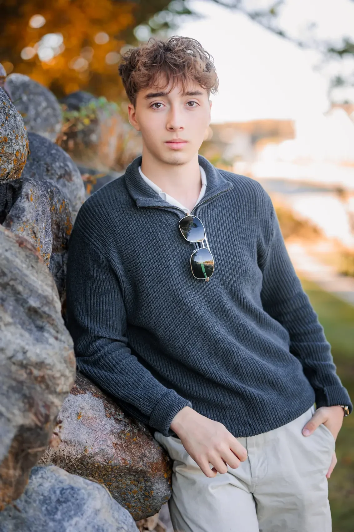 Senior boy with curly hair, wearing a dark blue sweater and white pants, leaning against a rocky wall with sunglasses hanging from his shirt. Fall trees in background.  Photo by Wendy Sorensen, a Des Moines senior photographer.