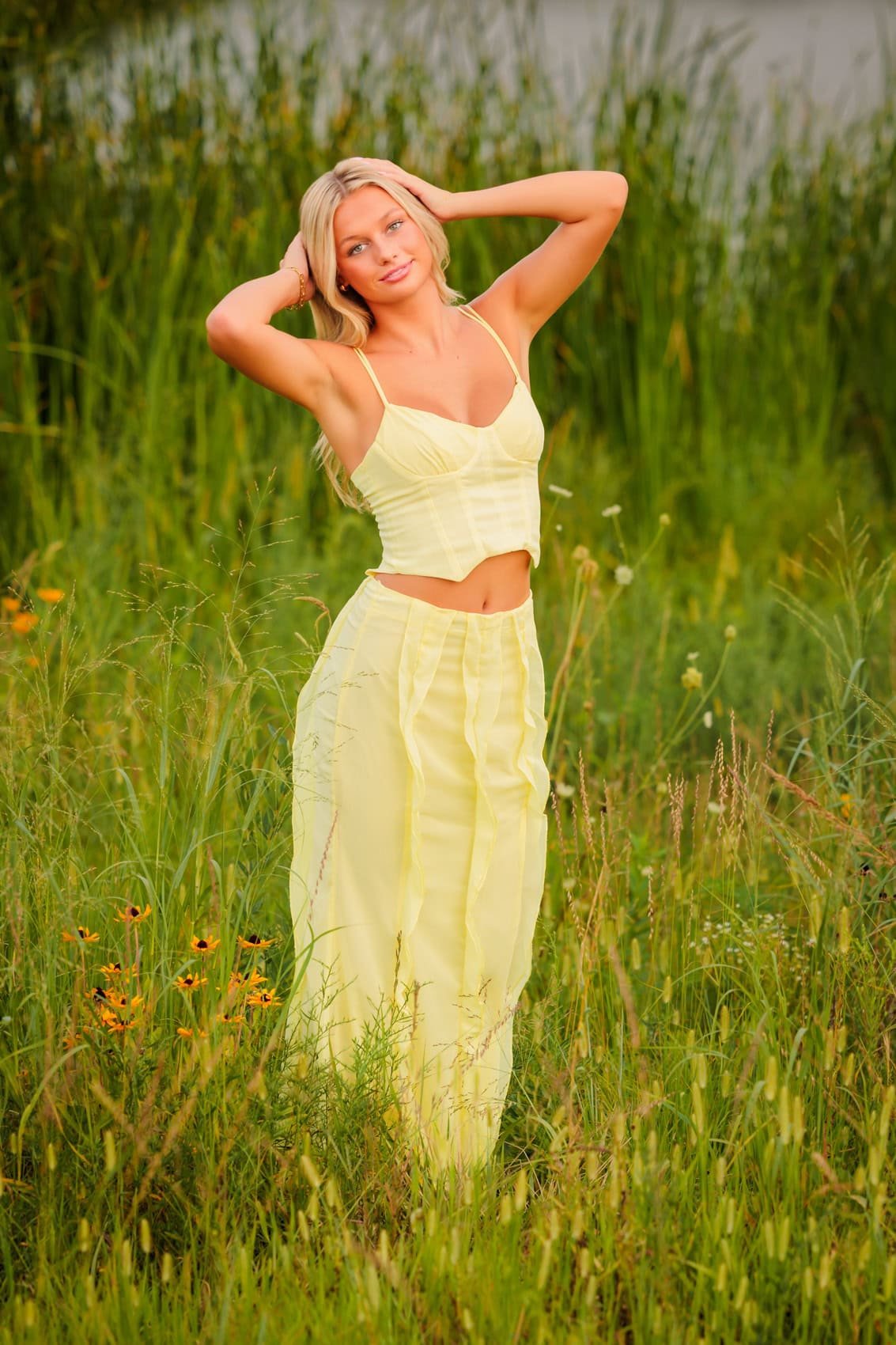 A young woman in a yellow outfit standing in a grassy field with wildflowers, posing with her hands behind her head.