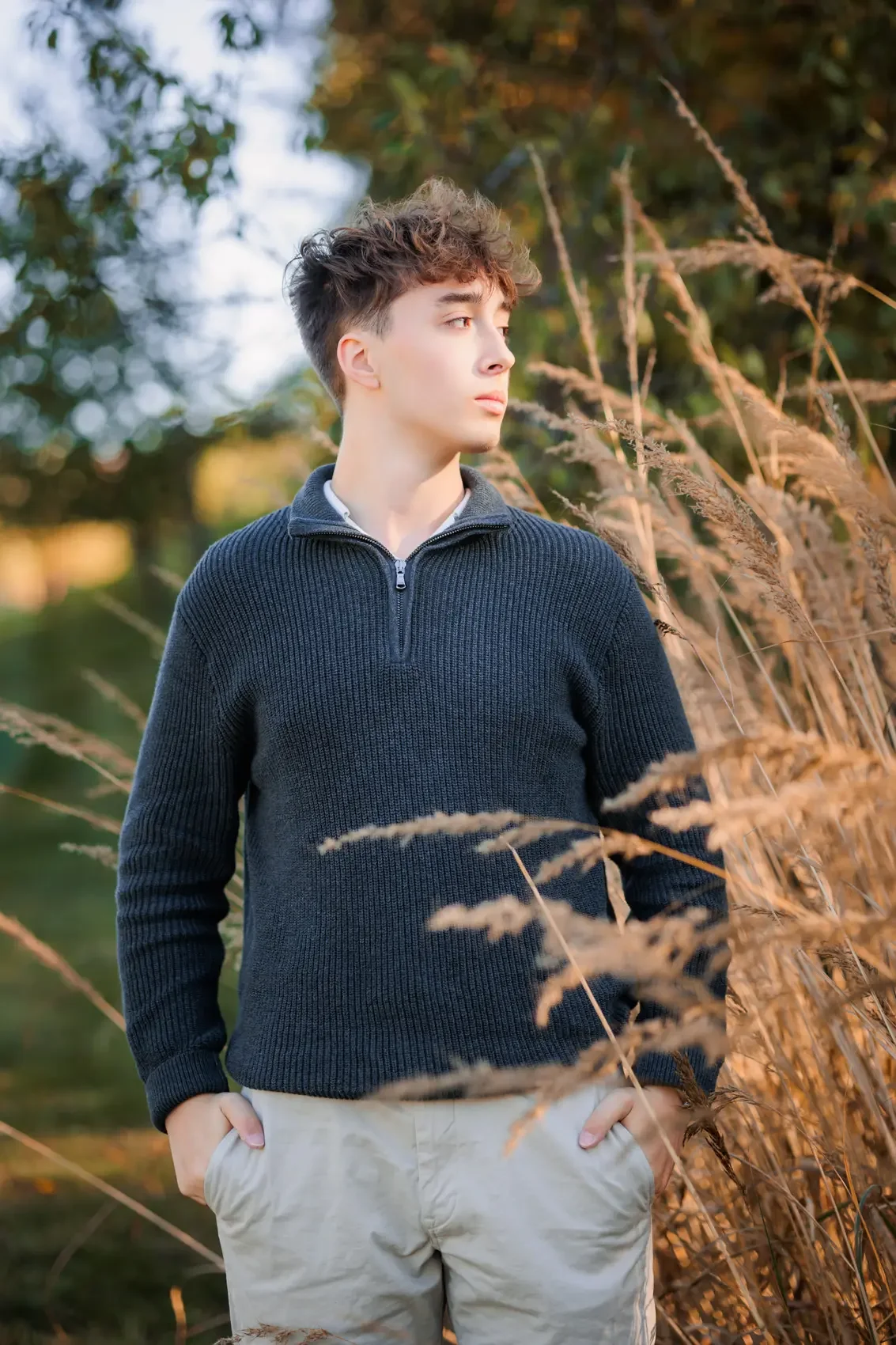 A young man with light skin and curly brown hair standing outdoors in a field of tall dry grass, looking to the side with a contemplative expression, wearing a navy blue sweater with a quarter zipper and beige pants.