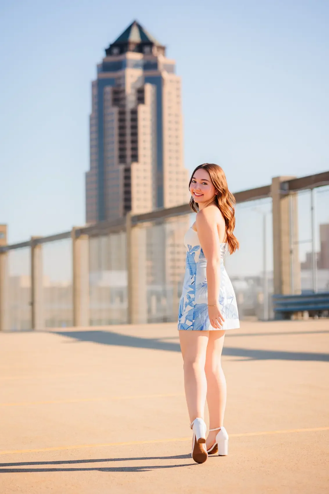 Senior girl with long brown hair, smiling and wearing a blue and white floral dress with high heels, standing on a parking garage rooftop in Des Moines with The Principle Building in the background on a sunny day.