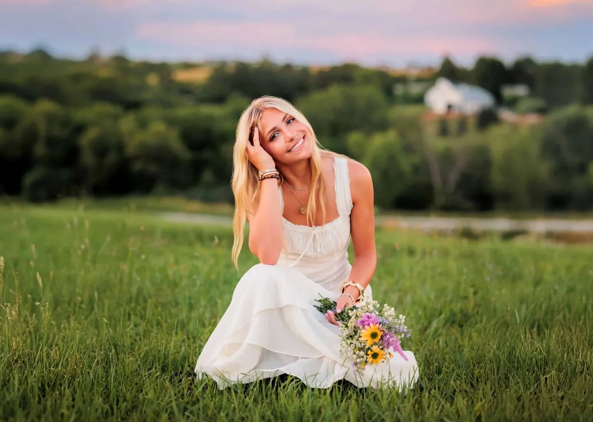 Senior girl with long blonde hair wearing a white dress and holding a bouquet of wildflowers, sitting on the grass.  Photo by Wendy Sorensen, a Des Moines senior photographer.