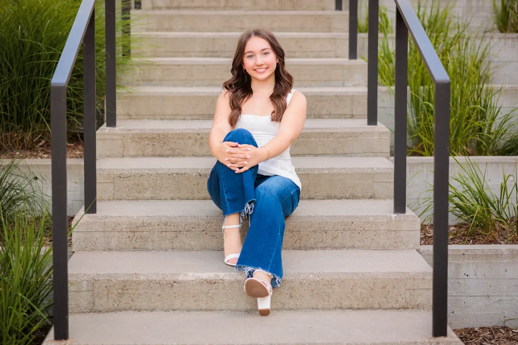 Senior girl with long brown hair, wearing a white sleeveless top, blue jeans, and white heels, sitting on outdoor concrete stairs with black metal handrails, surrounded by greenery. Photo by Des Moines senior photographer Wendy Sorensen.