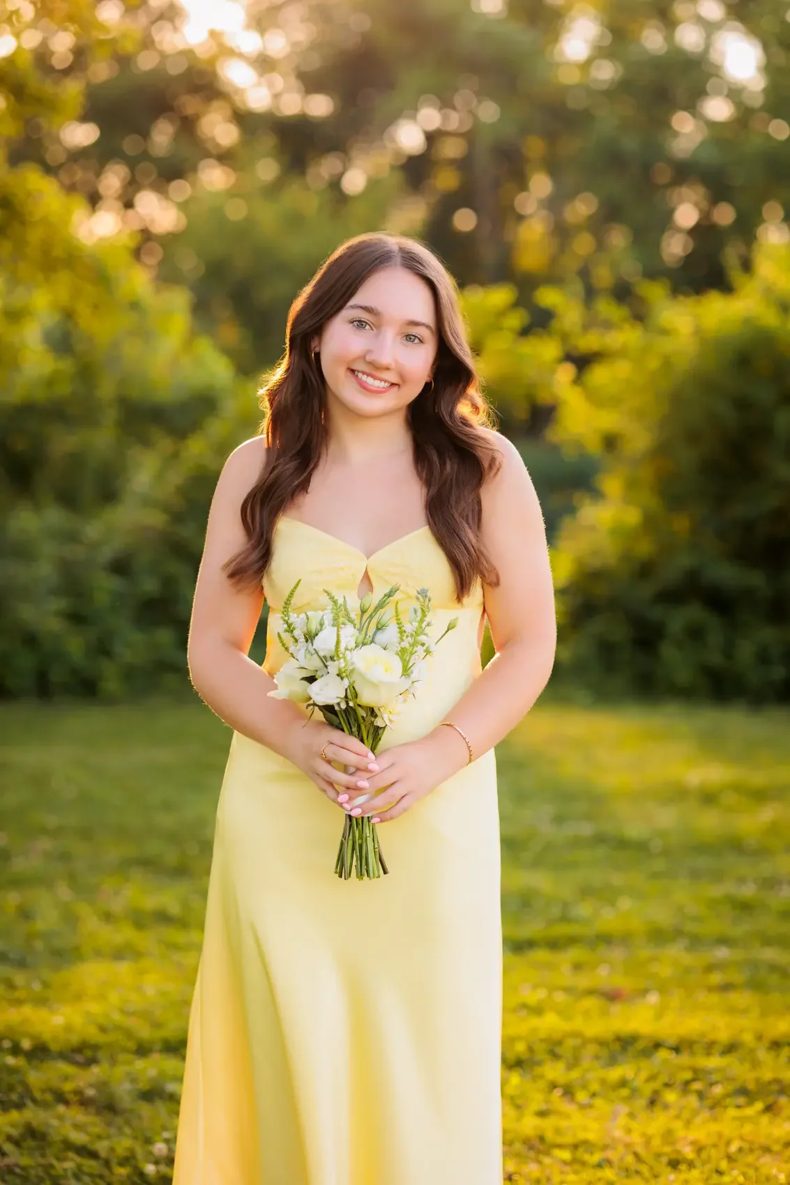 Senior girl in a yellow dress holding a bouquet of white flowers outdoors during sunset, smiling, at Water Works Park in Des Moines, Iowa.
