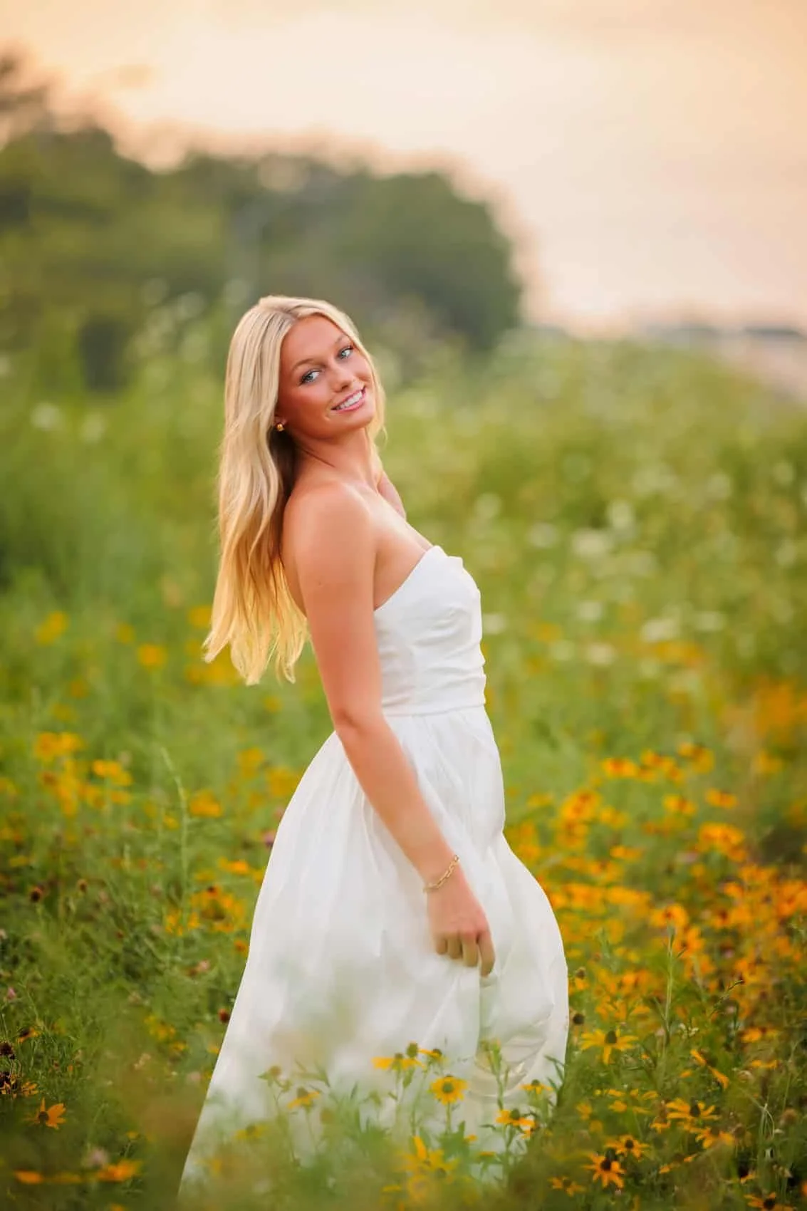 A young woman with long blonde hair wears a strapless white dress and stands in a field of yellow flowers at sunset, smiling and looking over her shoulder.