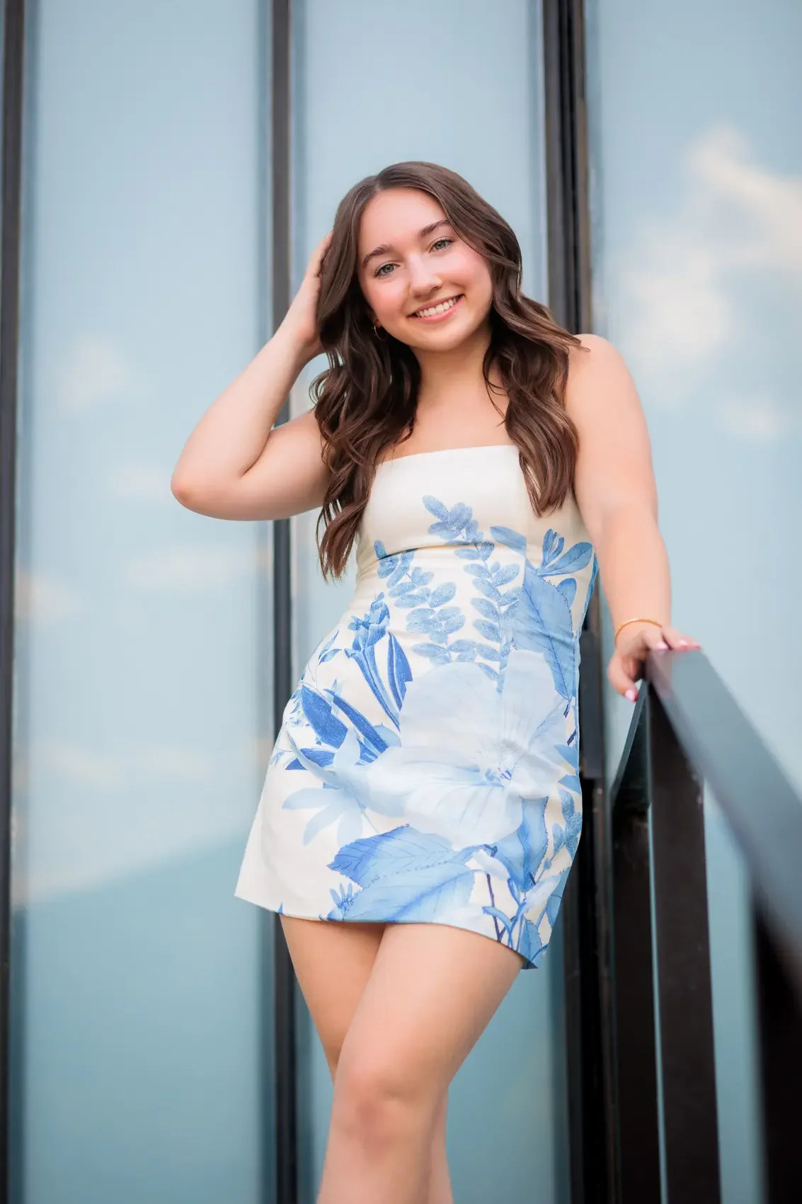 Senior girl with long brown hair, wearing a white dress with blue floral patterns, smiling and leaning on a railing outdoors, with a glass building and sky in the background in Des Moines, Iowa.