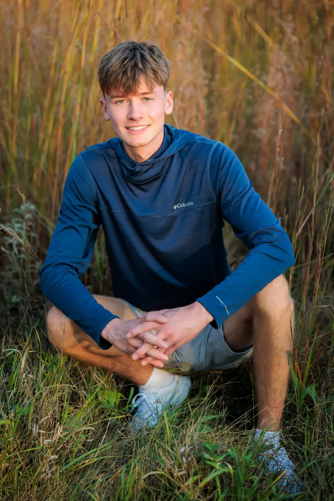Senior boy with short brown hair, blue eyes, wearing a blue Columbia long-sleeve shirt and gray shorts, squatting outdoors in a grassy area with tall grass and plants.