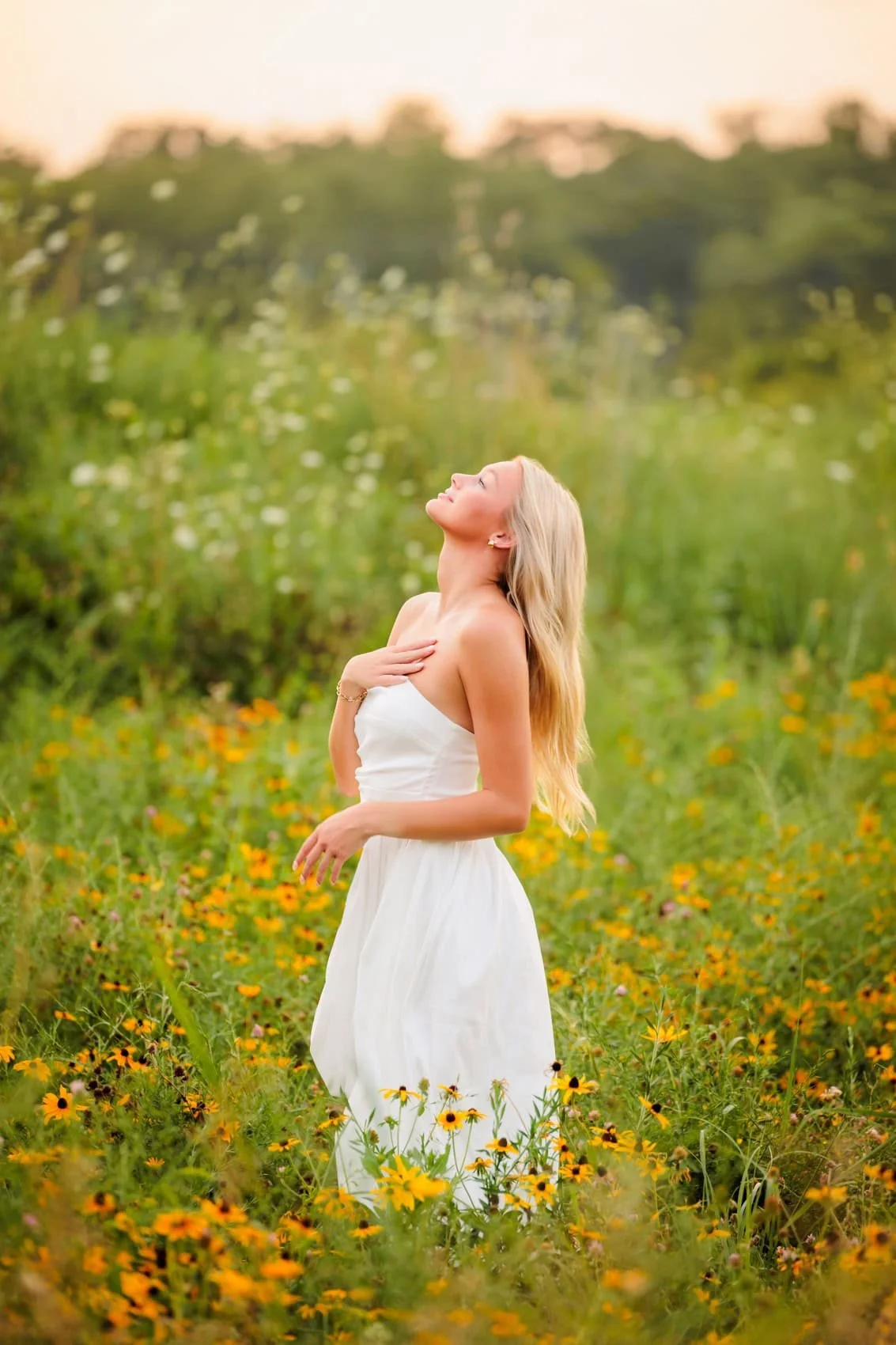 Senior girl with long blonde hair wearing a white dress in a field of yellow and orange flowers, looking up with her hand on her chest and a serene expression.