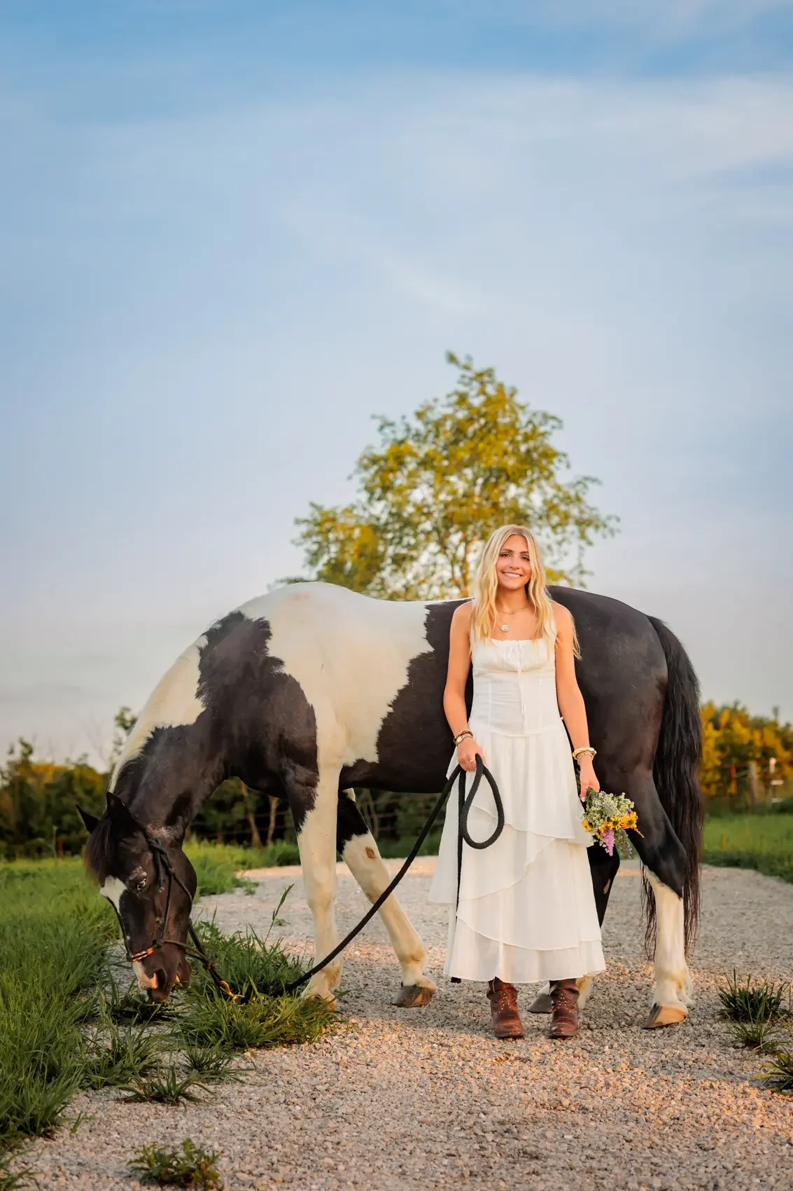 Senior girl in a white dress standing next to a black and white horse outdoors, holding a bouquet of flowers, with green grass and trees in the background under a blue sky.