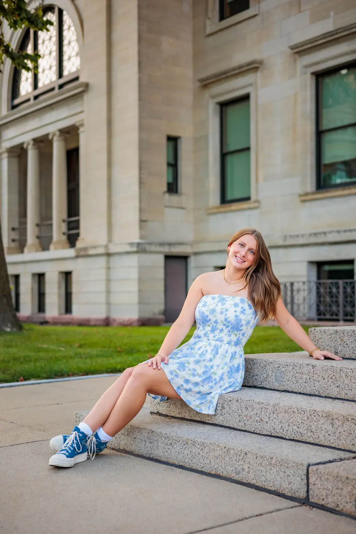 Senior girl sitting on steps outdoors, wearing a light blue floral dress and blue sneakers, smiling at the camera at the State Library of Iowa in downtown Des Moines.
