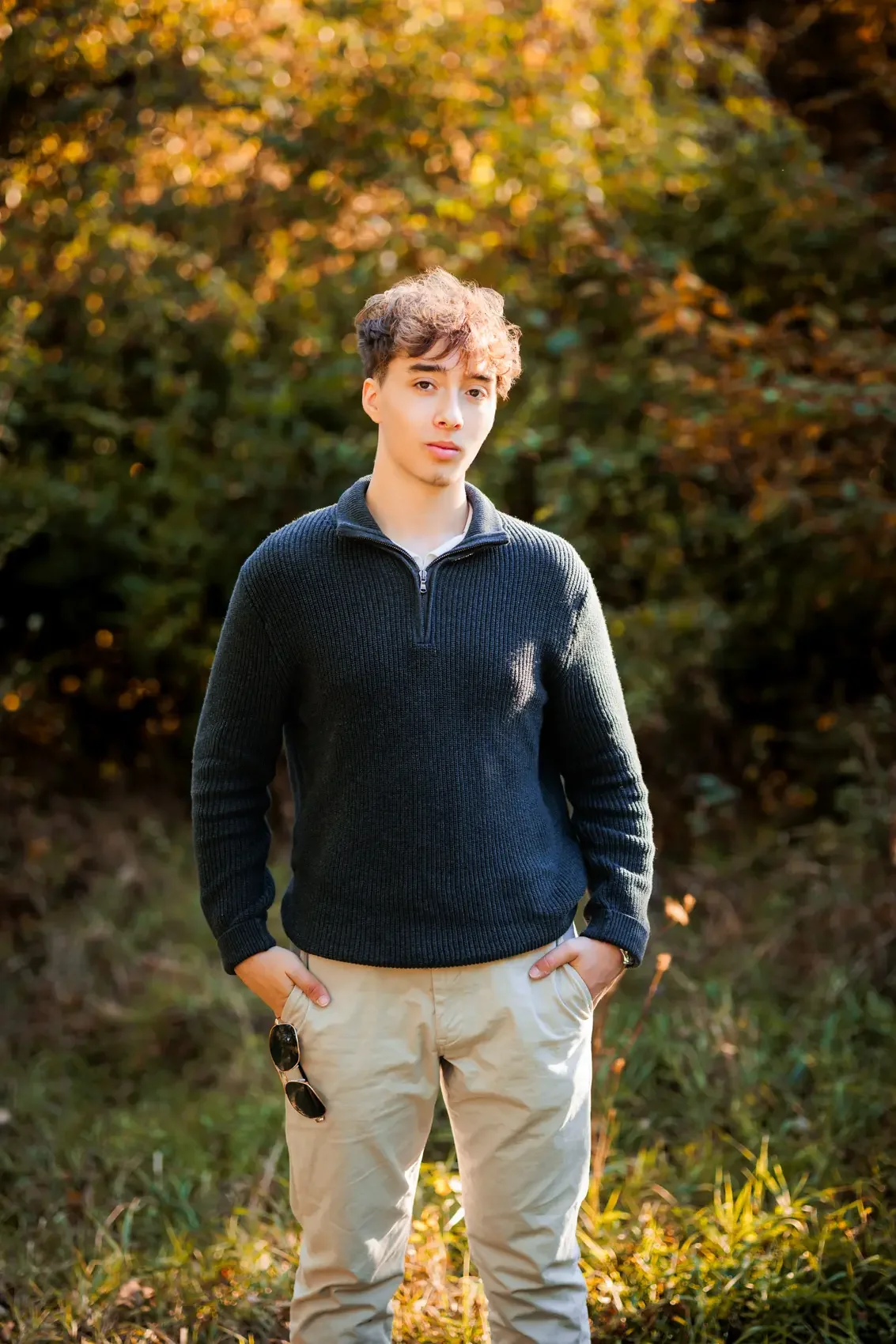 A young man with curly brown hair and light skin stands outdoors during autumn, wearing a black sweater and beige pants, with sunglasses hanging from his pocket, in front of a background of trees with orange and green leaves.