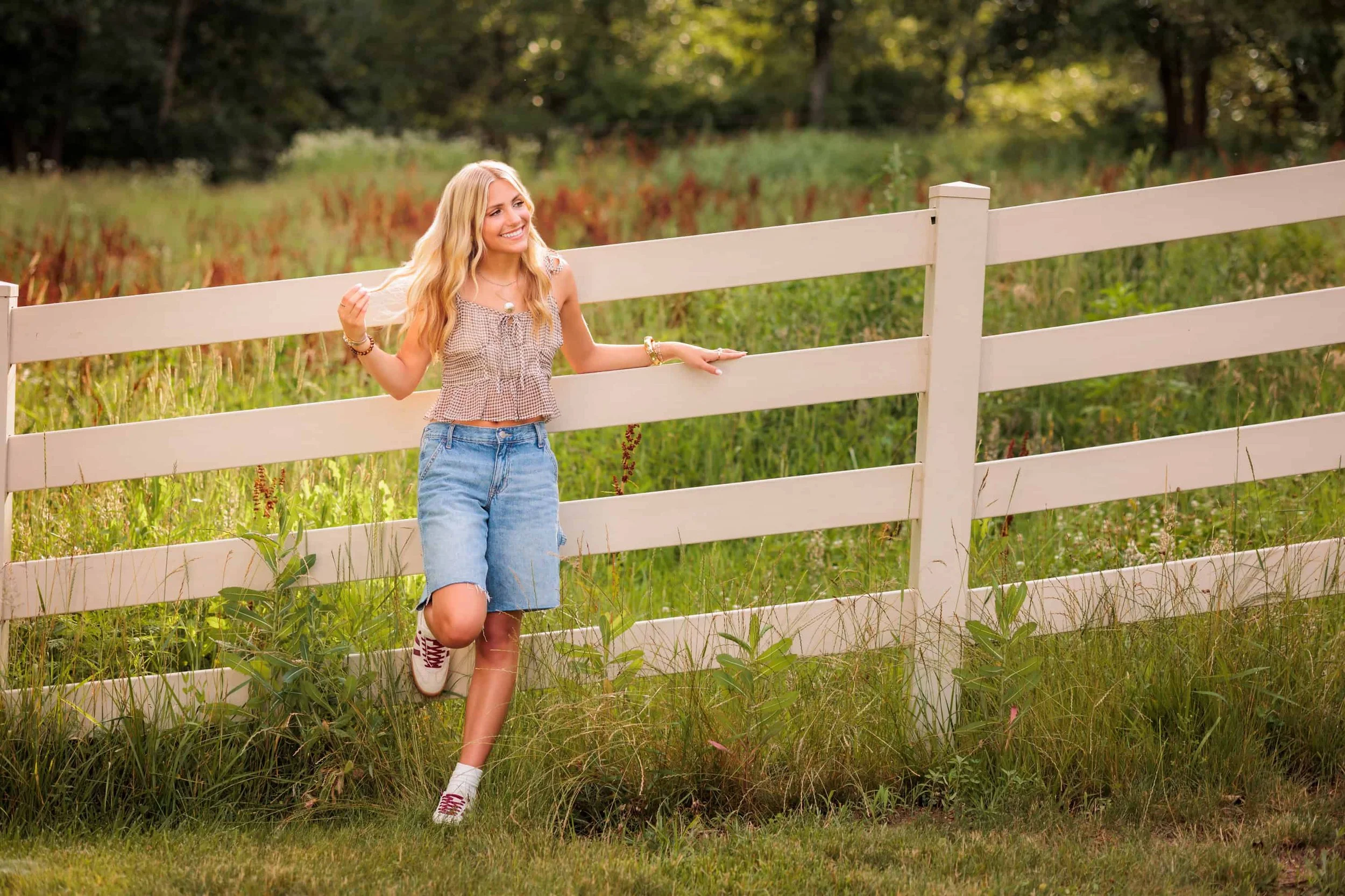 Senior girl with long blonde hair leaning on a fence out in the country, playing with her hair and looking off to the side.