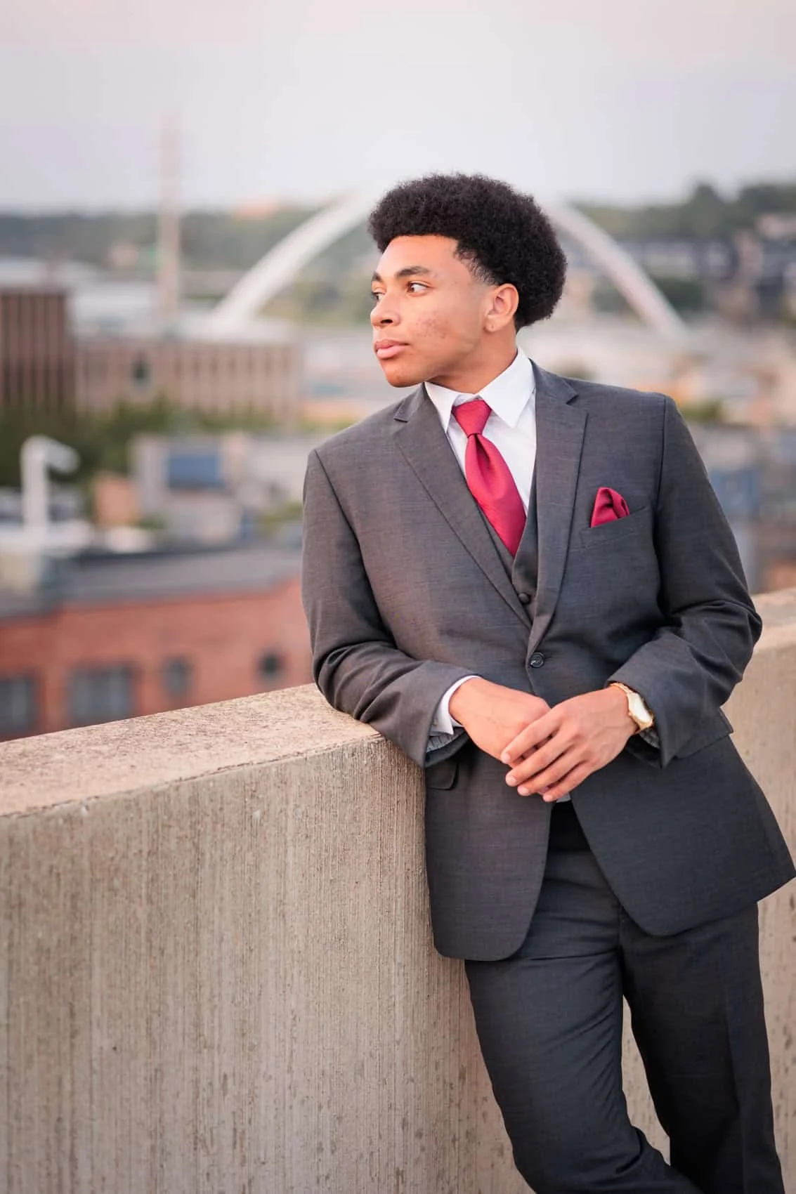 High school senior guy in a gray suit and red tie leaning against a concrete railing on a rooftop with a cityscape in the background, including a large arch bridge.