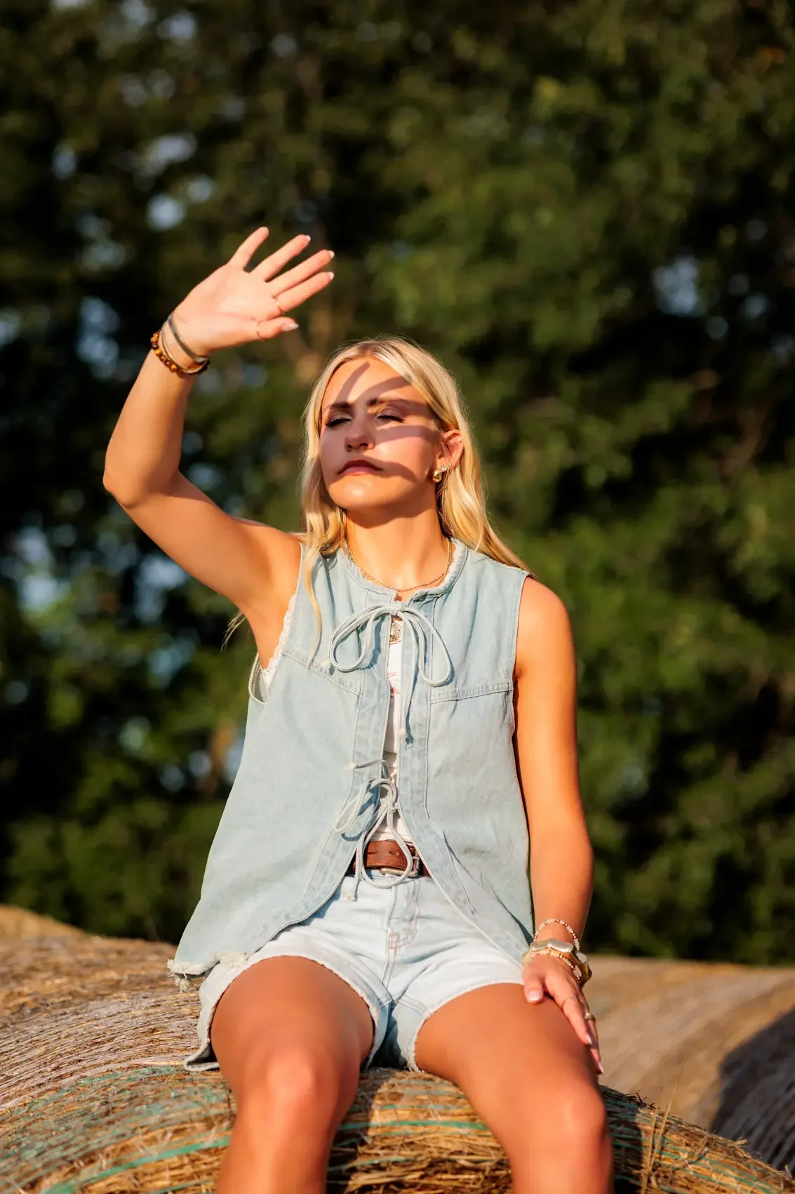 Senior girl sitting on a large log outdoors, shielding her eyes from the sunlight with her hand.