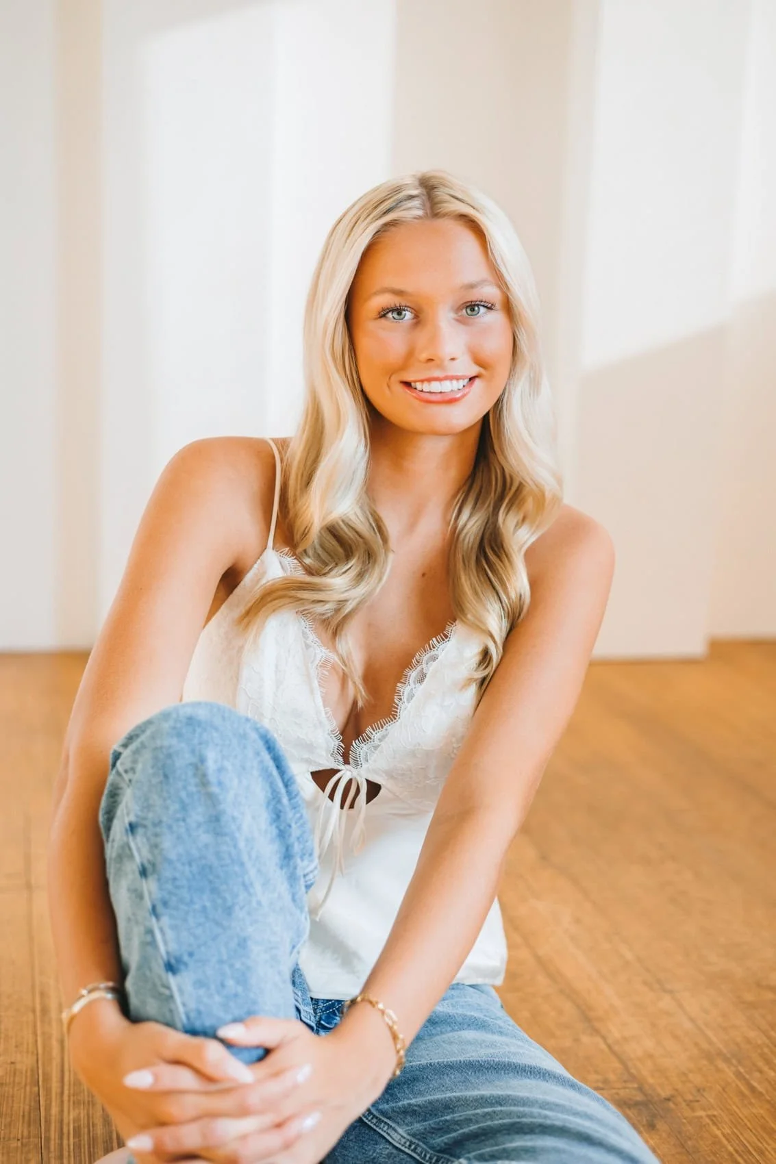Senior girl with long blonde hair and blue eyes sitting on the floor, smiling at the camera, wearing a white sleeveless top and blue jeans.
