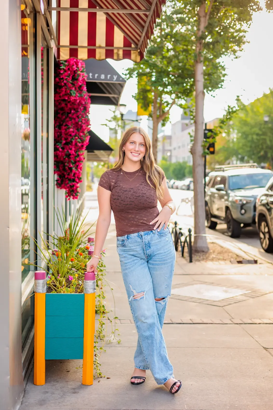A young woman with long wavy hair smiling and walking on the sidewalk beside a storefront with colorful flower pots and a striped awning, with trees and parked cars in the background.