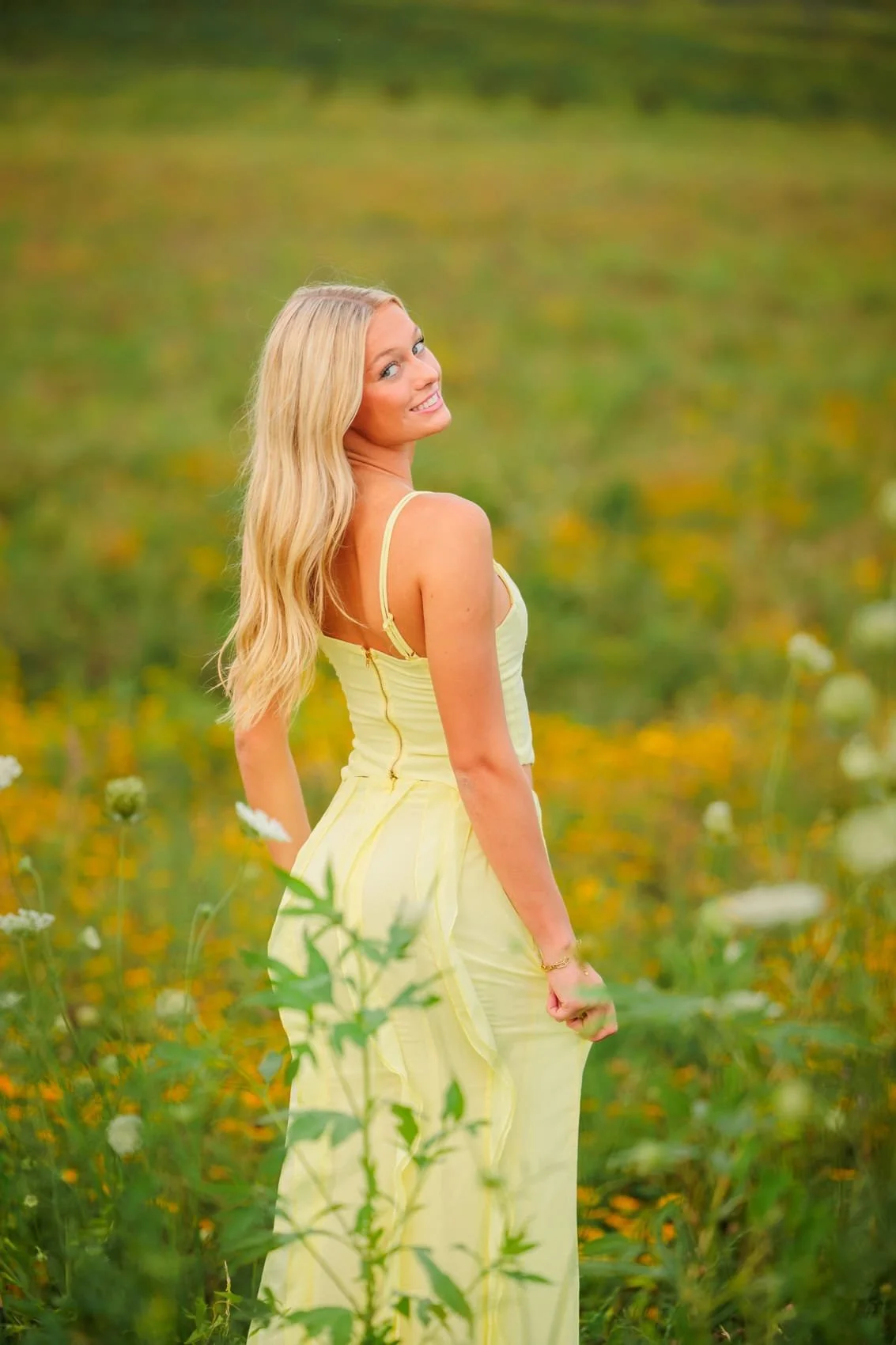 Senior girl with long blonde hair wearing a light yellow dress, standing in a green and yellow wildflower field, smiling and looking over her shoulder. Photo by Wendy Sorensen, a Des Moines senior photographer.