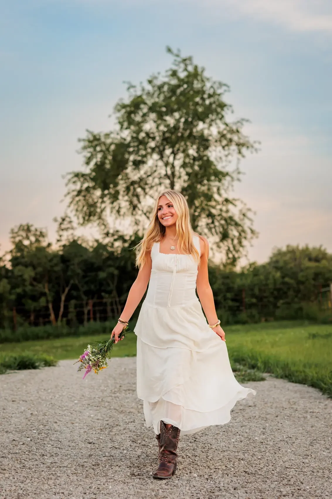 Senior girl in a white dress and cowboy boots walking on a gravel path, holding a small bouquet of flowers, with a large tree and green landscape in the background.