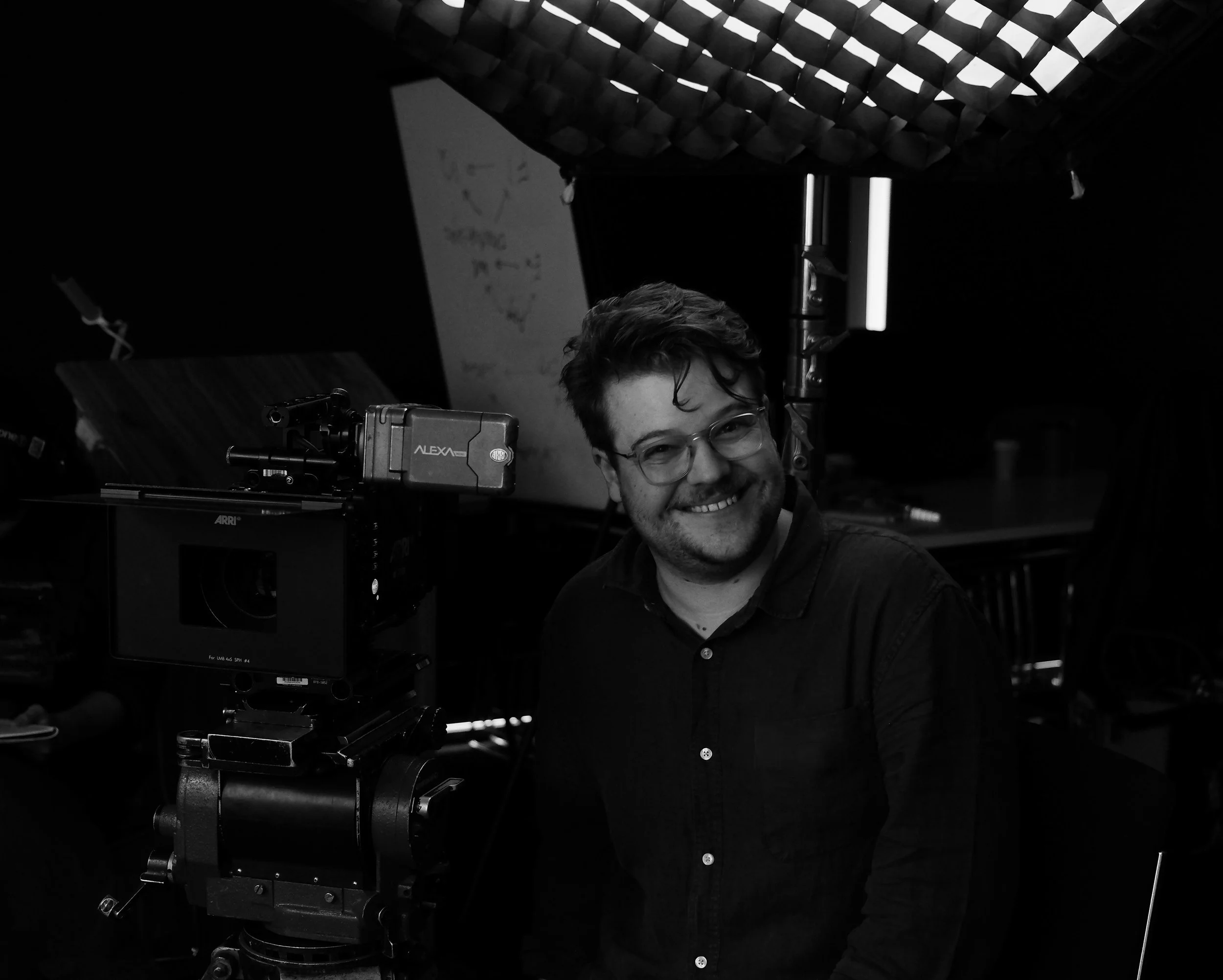 Black and white photo of a man with glasses smiling behind film production equipment, with a patterned ceiling above.