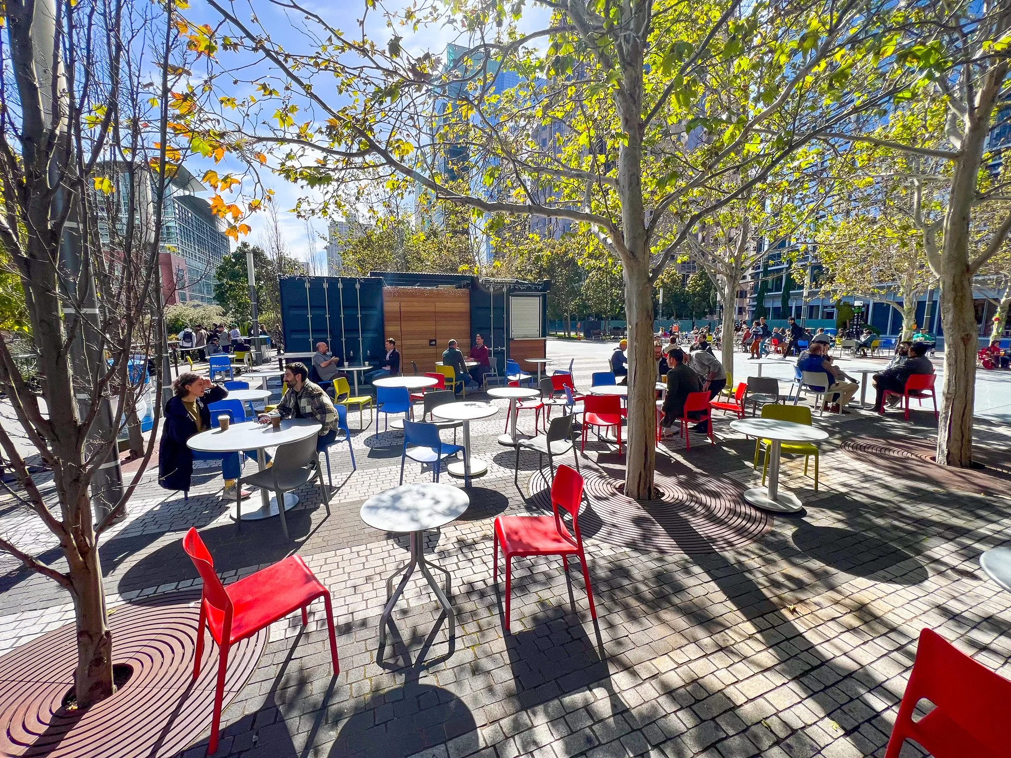 Outdoor patio area with colorful chairs and tables shaded by trees, with people sitting and chatting on a sunny day in an urban setting.