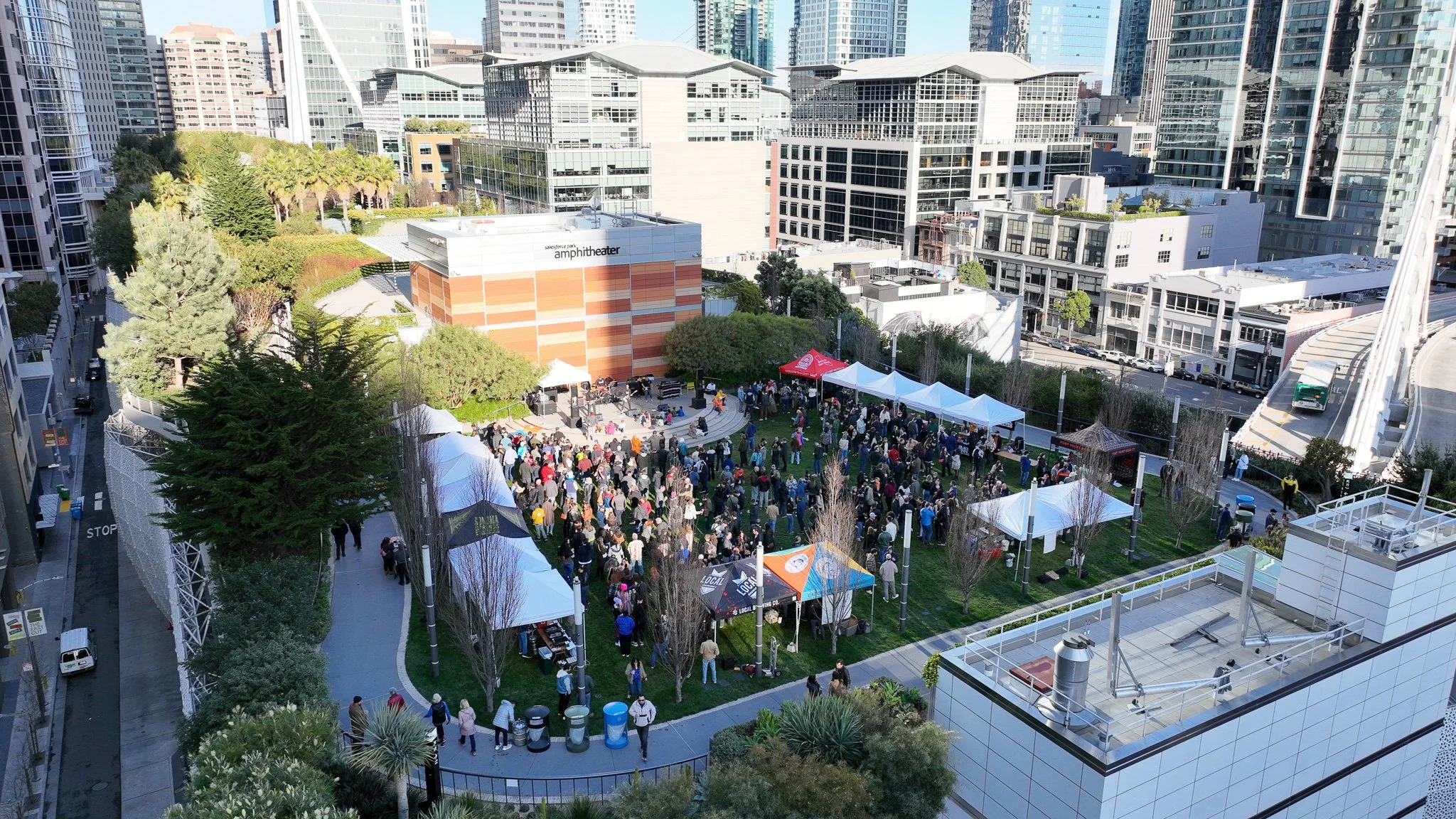 Crowd gathered in an outdoor urban park with tents and a stage, surrounded by tall modern buildings.