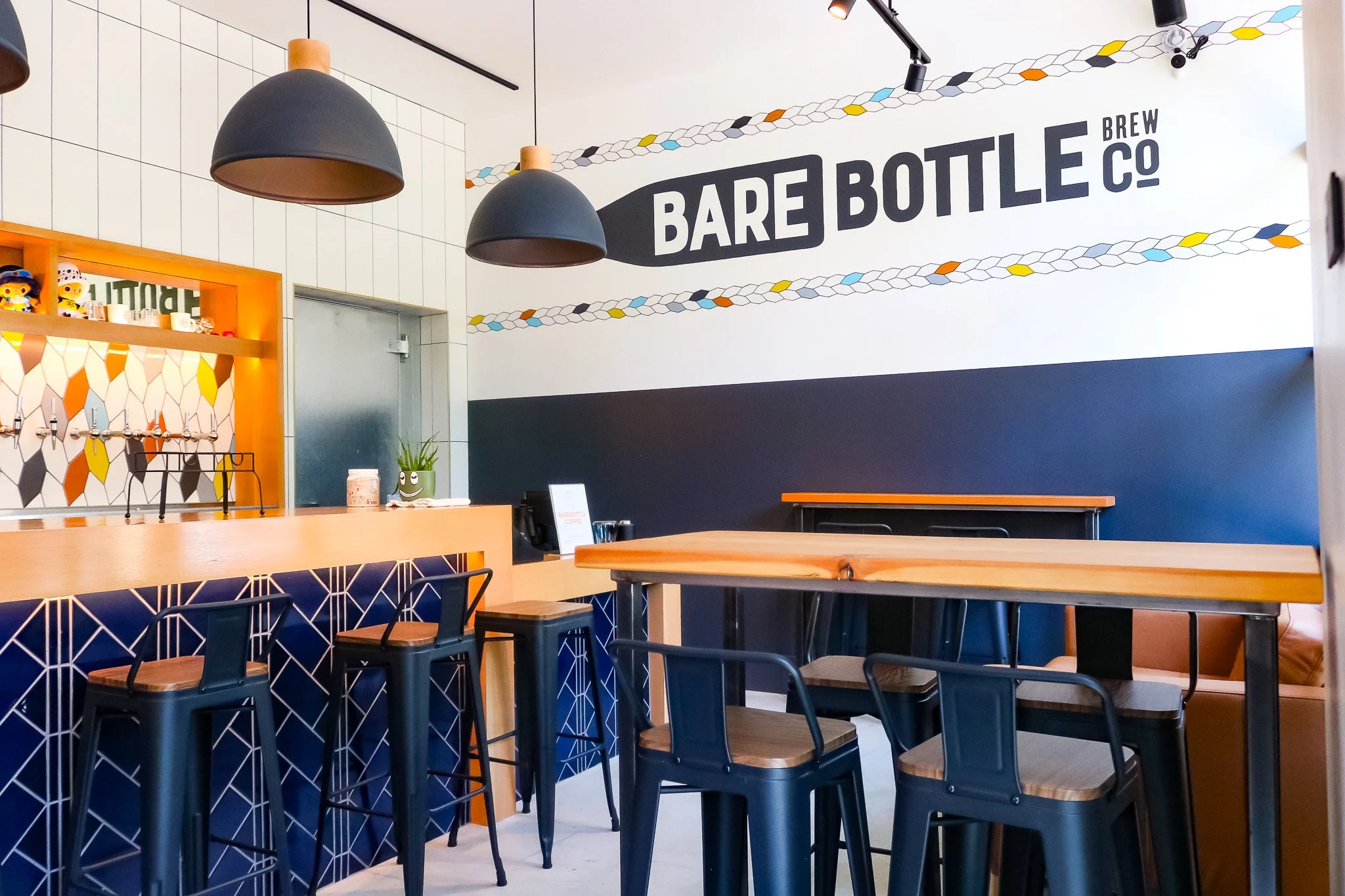 Interior of Barebottle Menlo Park with wooden tables and benches, large windows, pendant lights, and a tiled wall with a sign.