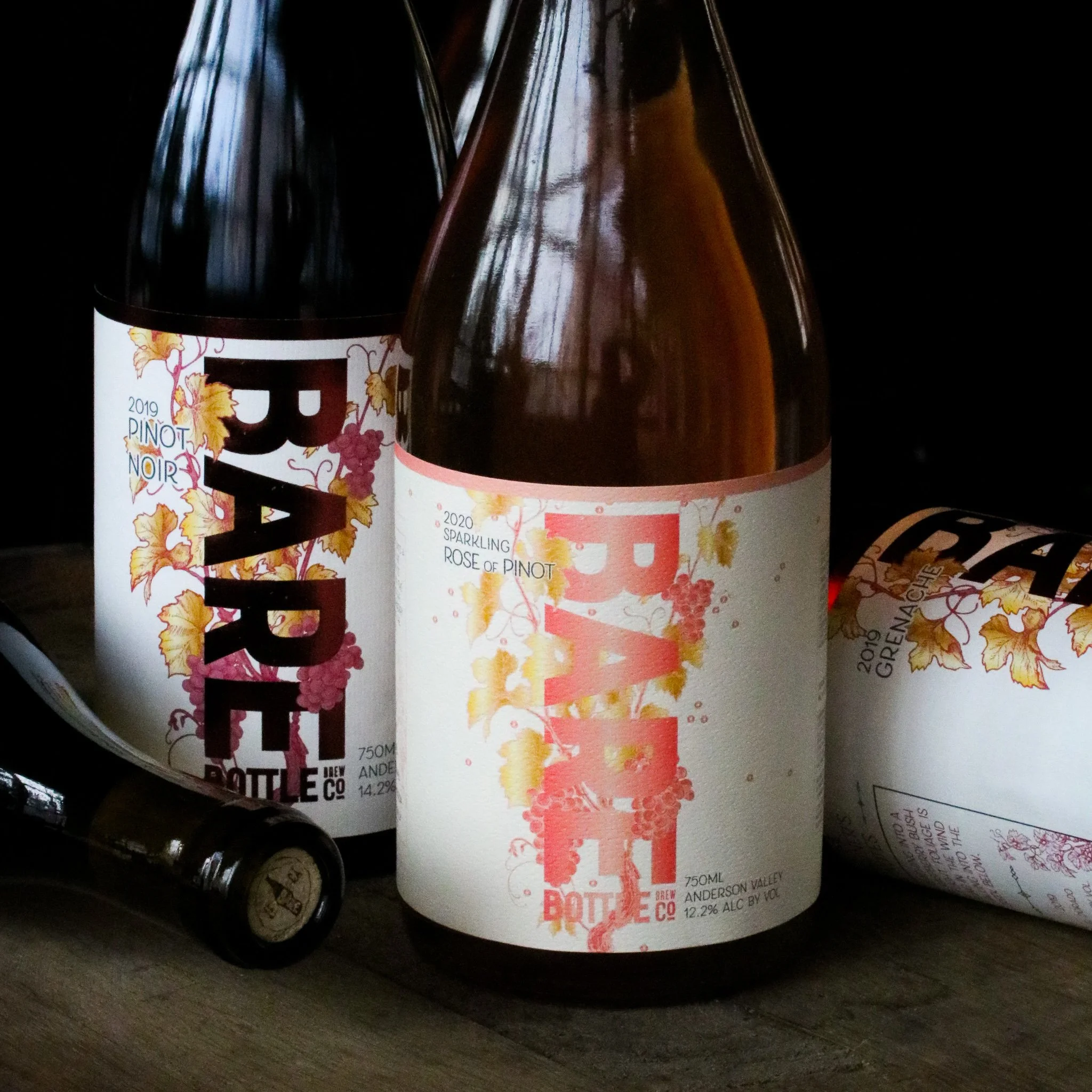 Three bottles of wine with colorful labels and a rolled paper label, one of which is lying on its side. The bottles are placed on a wooden surface against a dark background.