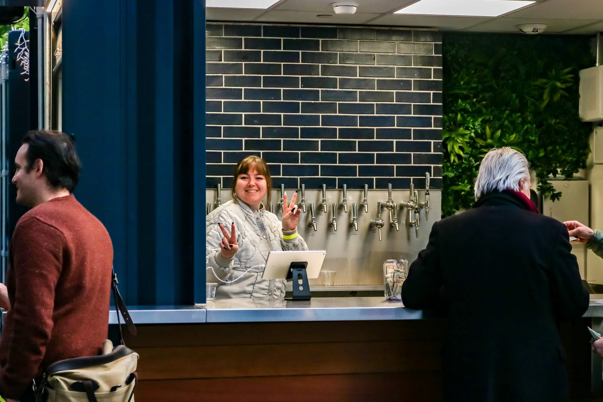 A woman behind a bar counter smiling and making peace signs with both hands. She has light brown hair and is wearing a gray jacket. The bar wall is equipped with multiple taps and has a dark brick design. Two men are in front of the counter, one on the left in a brown sweater and tan bag, the other on the right with white hair and a black jacket. There are some glasses and a tablet on the counter.