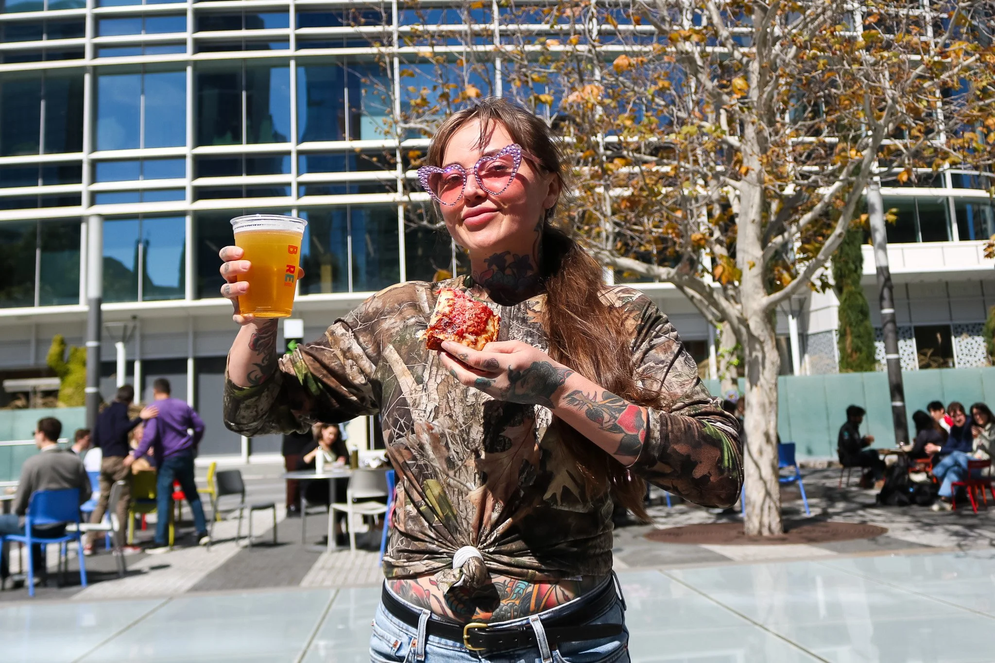 A woman with tattoos wearing pink heart-shaped sunglasses and a camouflage long-sleeve shirt, tied at the waist, standing outdoors holding a cup of beer in one hand and a slice of pizza in the other. She is in a public space with people, tables, and a building with glass windows behind her, and a tree with autumn leaves.
