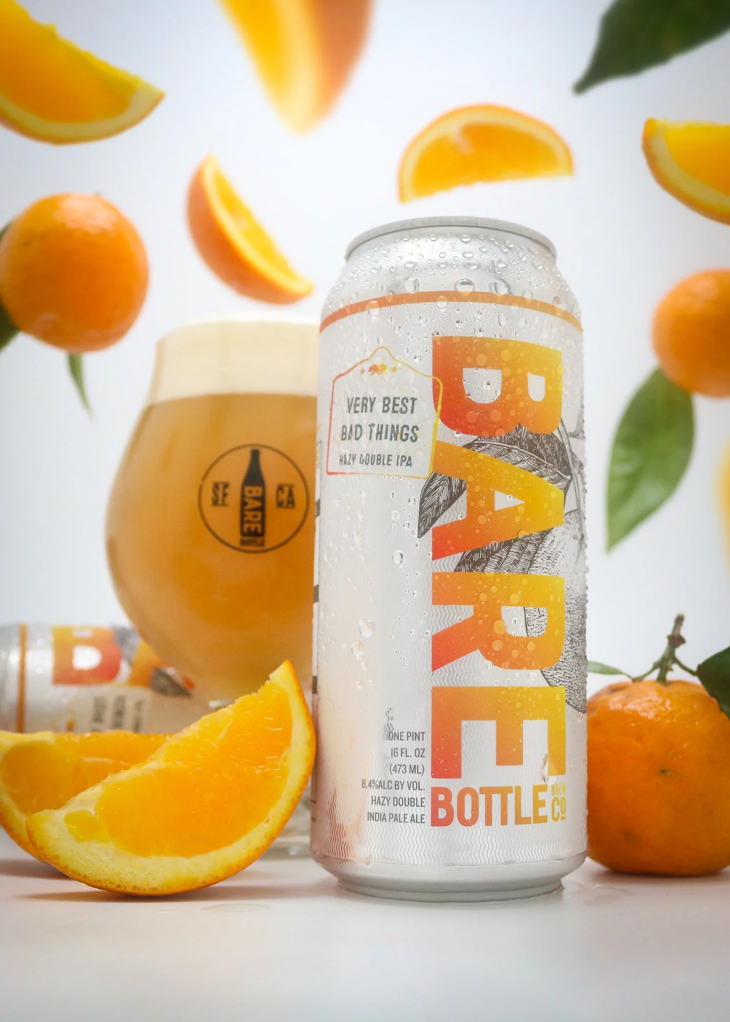 A can of Orange Bottle beer with citrus slices and oranges around it, in front of a glass of beer with a logo, and whole oranges and green leaves in the background.