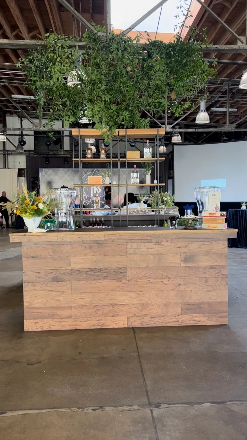 Wooden bar counter with decorative shelving above, decorated with flowers and bottles, in an indoor event space with concrete floor, ceiling with visible beams, and large green plants overhead.