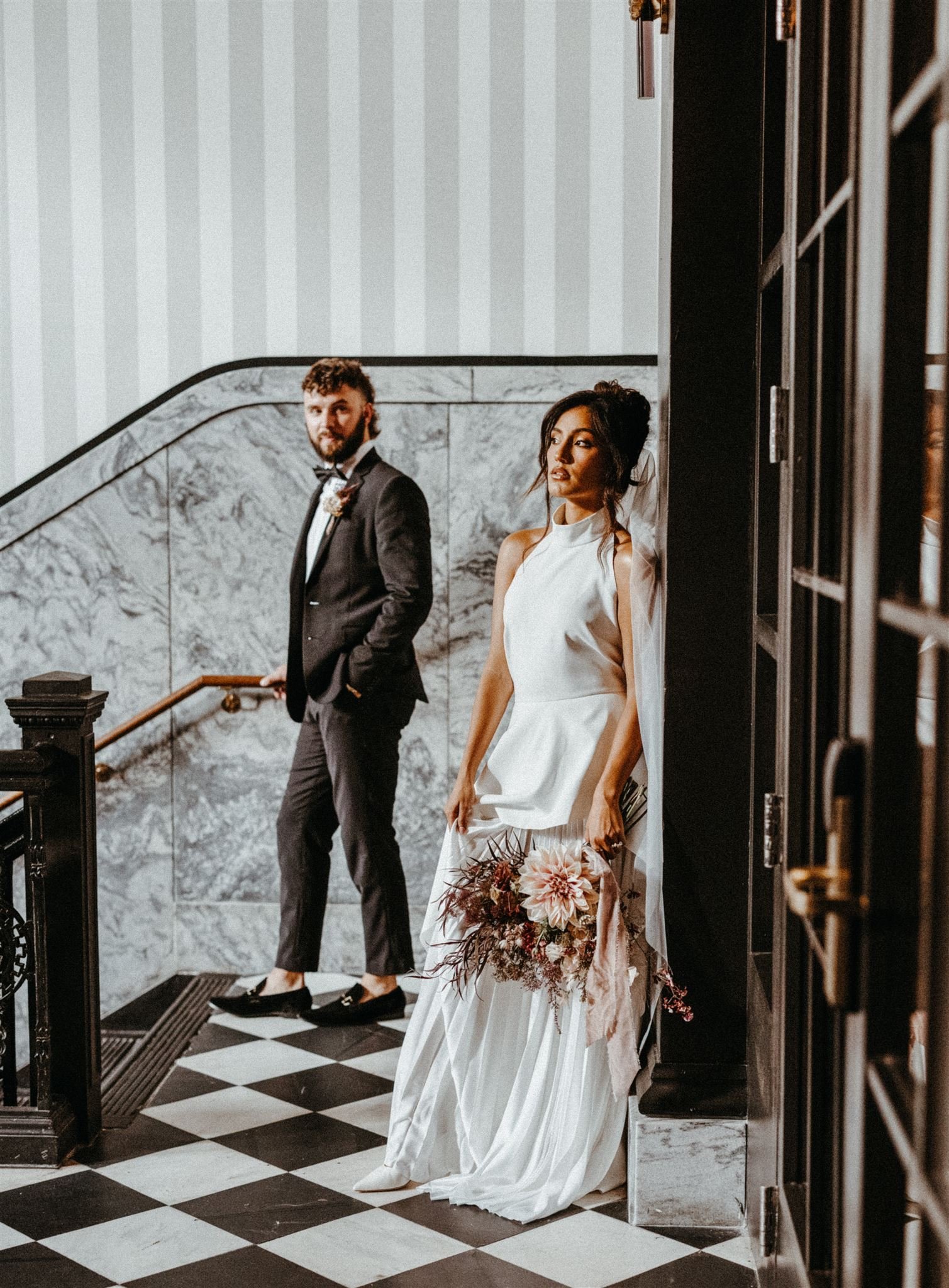 A bride in a white wedding gown holding a bouquet stands against a wall, with a groom in a black tuxedo with a white shirt and tie standing in the background, in an elegant indoor setting with black and white checkered flooring and marble walls.