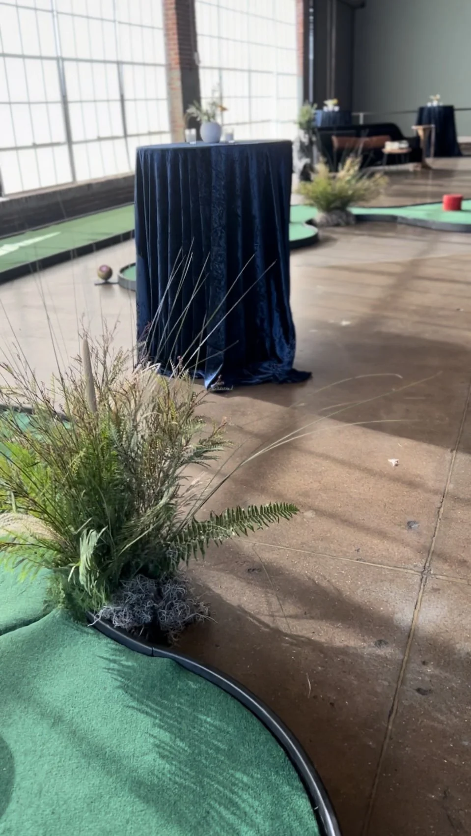Indoor miniature golf course with plants and a high-top table covered in dark blue fabric