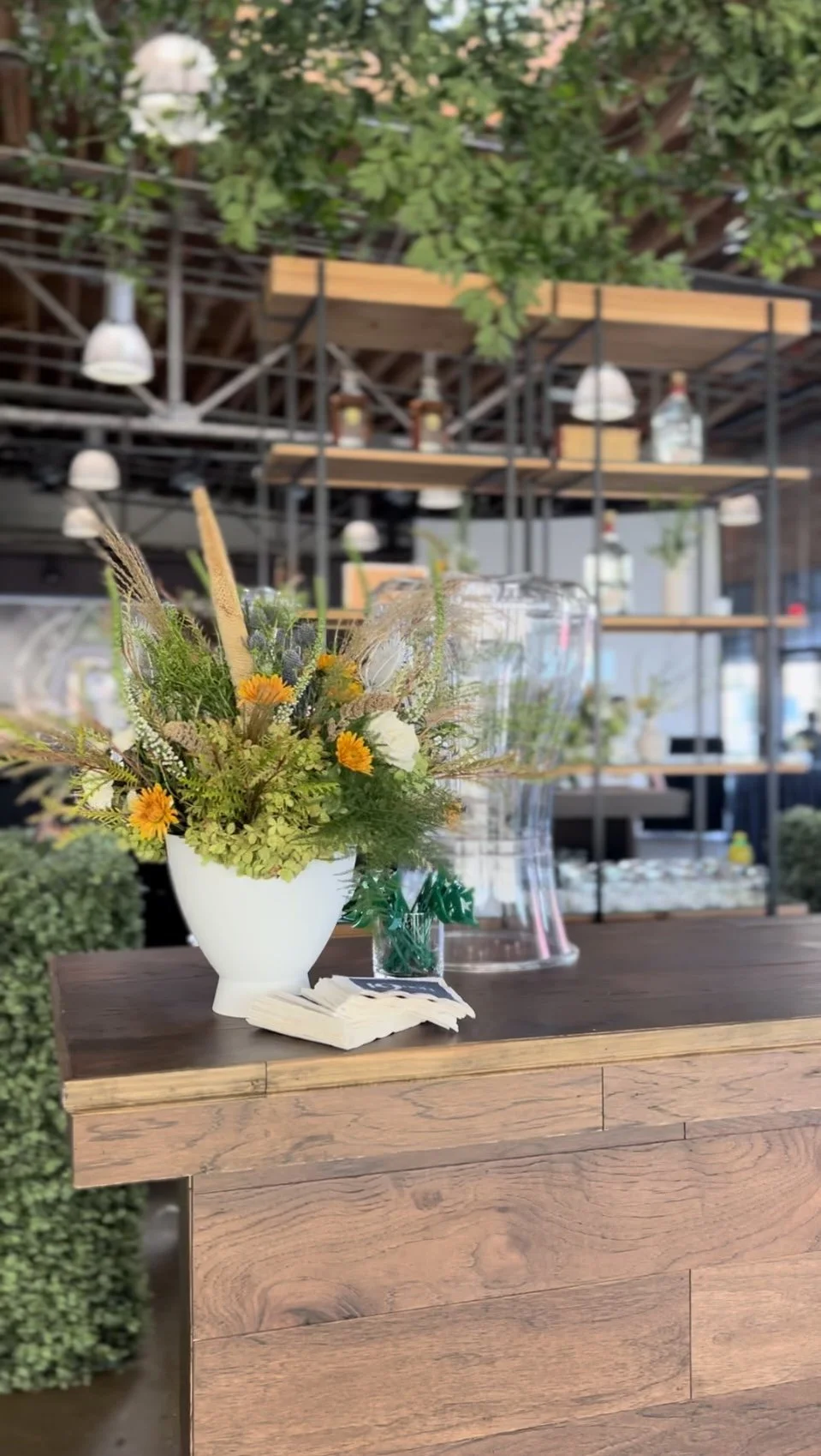 A floral centerpiece with yellow and white flowers on a wooden counter in a modern, industrial-style restaurant or cafe setting.