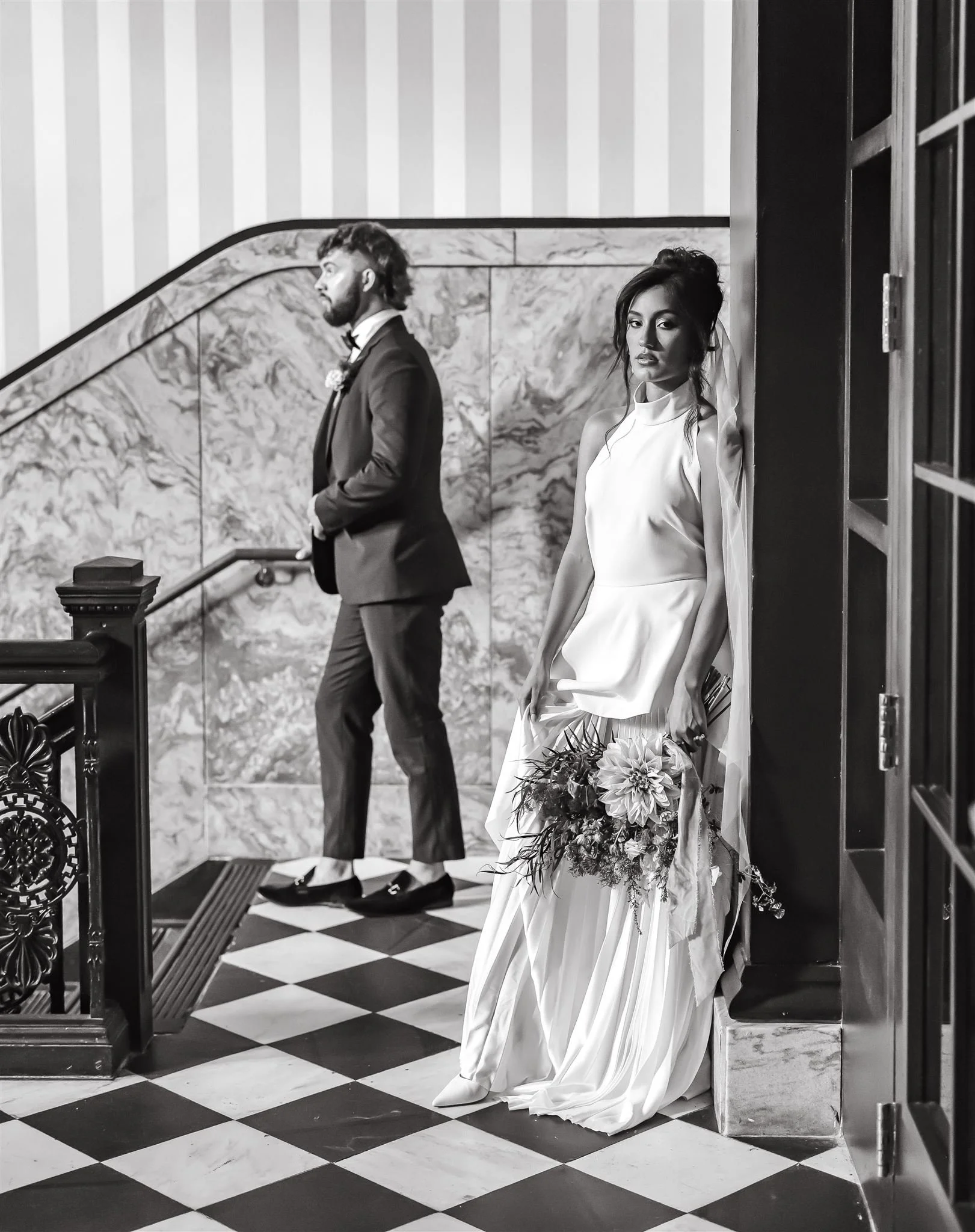 Black and white photo of a bride in a white wedding dress standing against a wall holding a bouquet, looking at camera. A groom in a tuxedo with a boutonnière stands in the background on stairs, facing sideways.