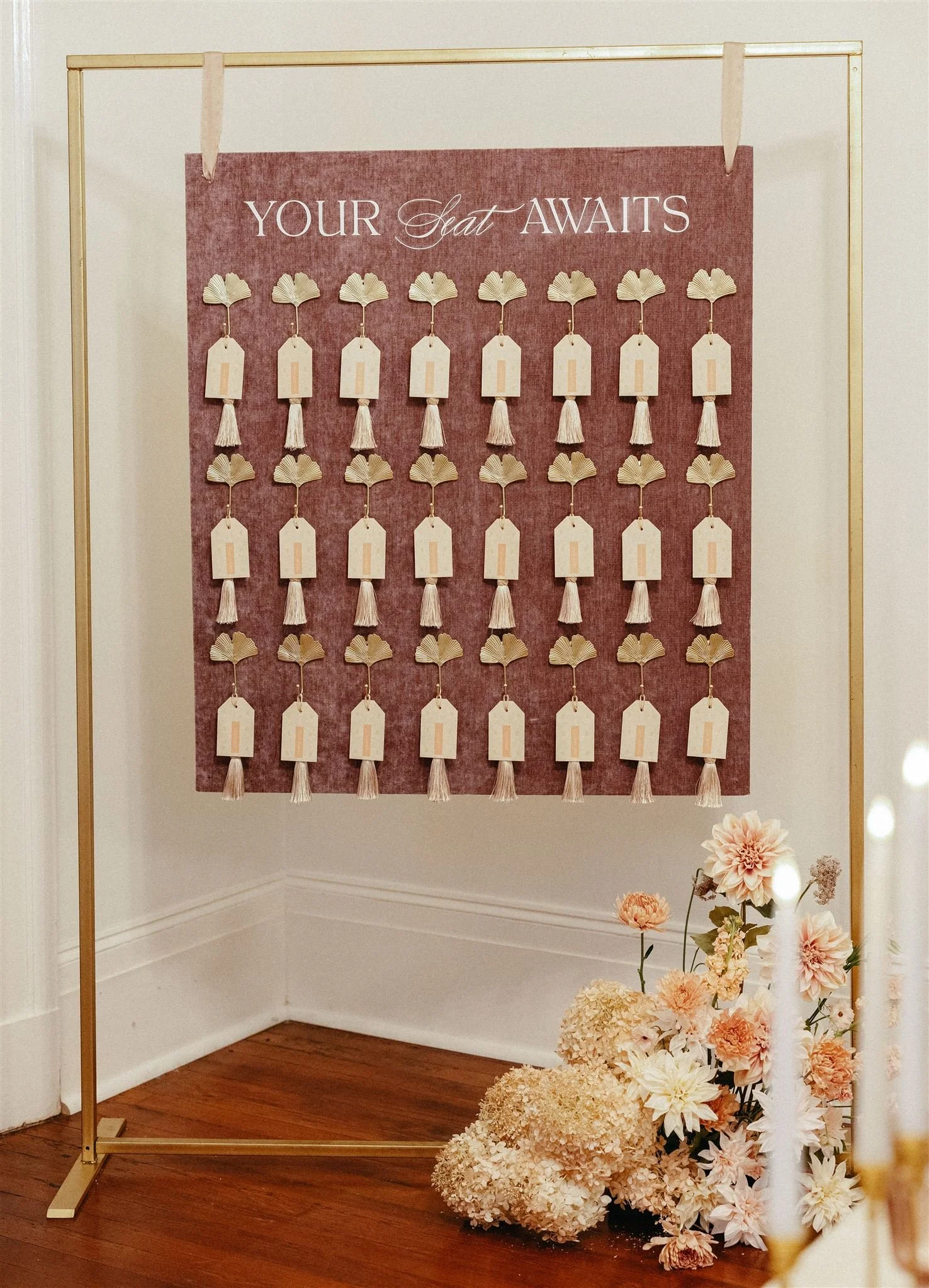A pink and gold display with small tags and tassels arranged in rows, labeled 'Your Seat Awaits,' next to a bouquet of pink and white flowers on a wooden floor.