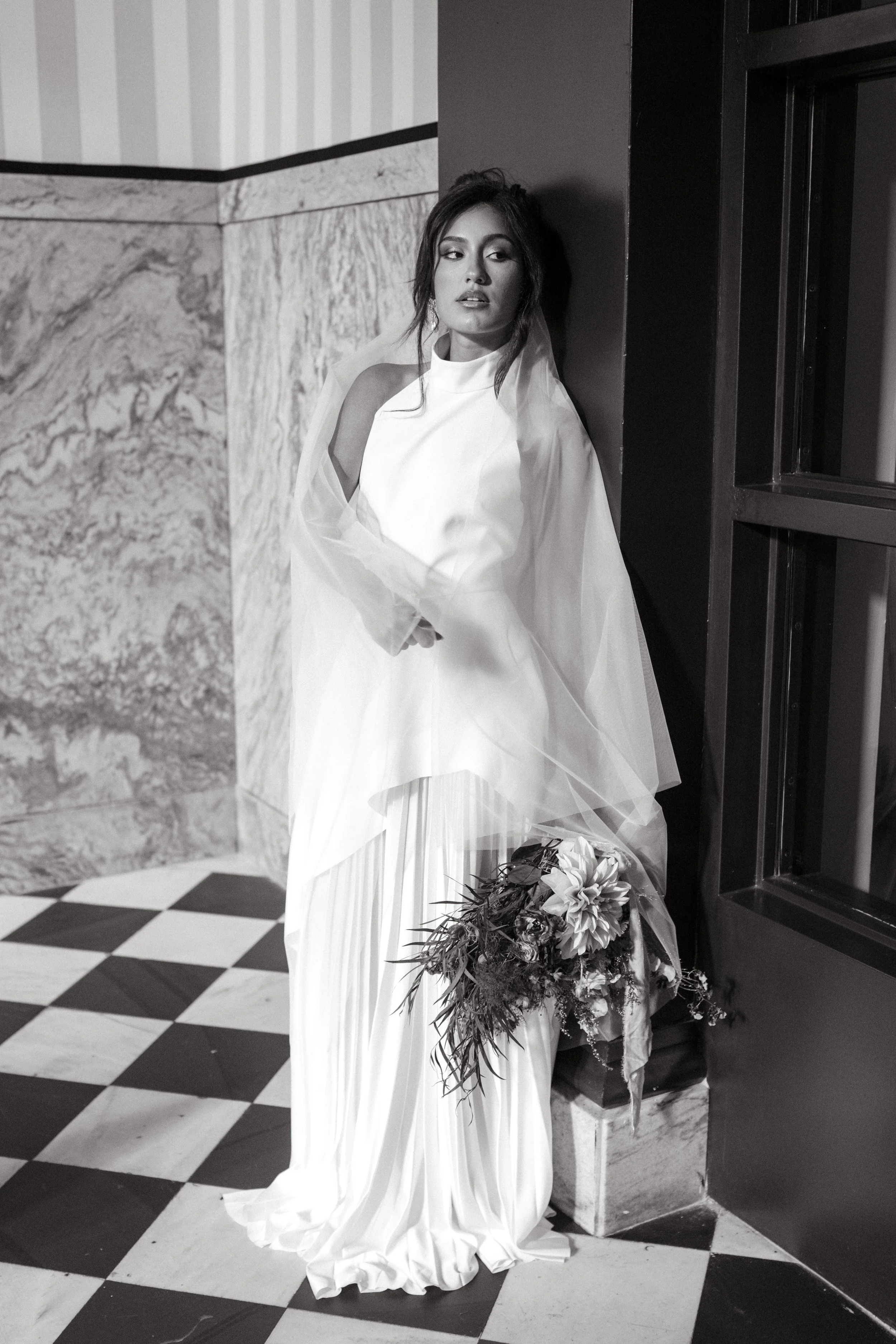 A woman in a white dress with a sheer cape, holding a bouquet of flowers, leaning against the corner of a marble wall in an indoor setting.