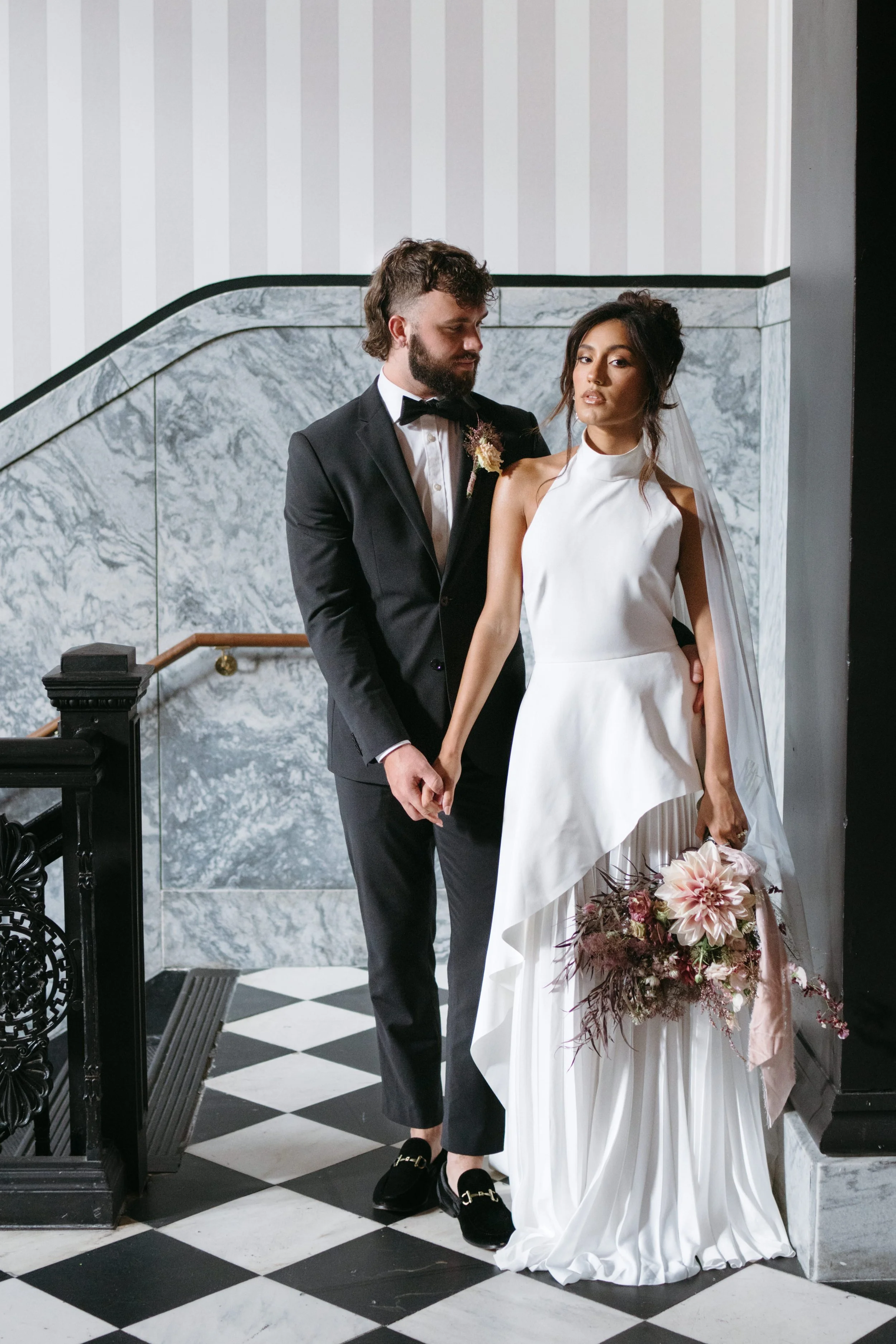 A bride and groom standing together inside a building, holding hands, with the bride holding a bouquet of flowers. The groom is dressed in a black tuxedo and tie, and the bride is wearing a white wedding dress with a high neckline and veil.
